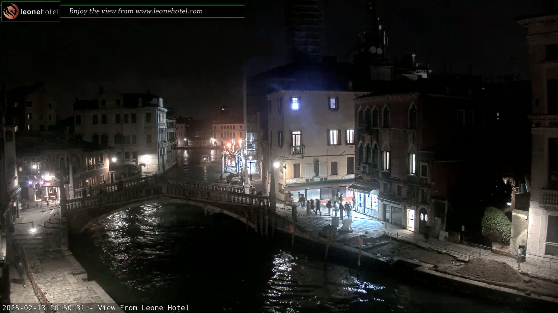 A nighttime view of a canal in Venice, Italy, shows a stone bridge over dark water, with buildings lining the canal banks and a few pedestrians visible.