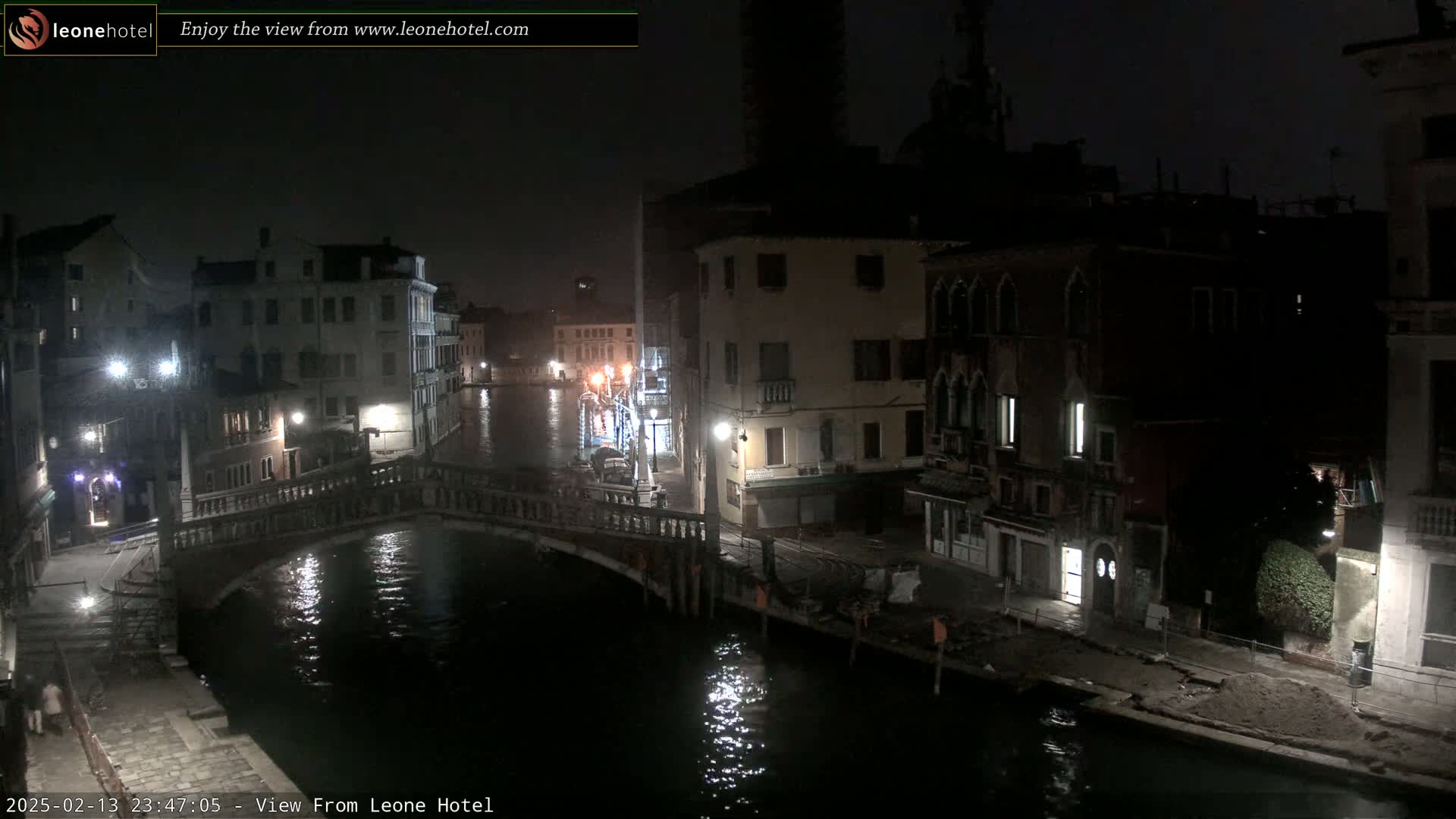 A nighttime view of a canal in Venice, Italy, shows dark buildings lining the waterway and a bridge spanning the canal.