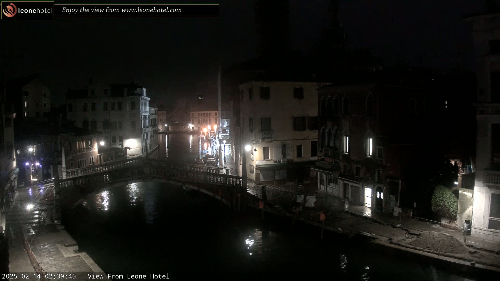 A nighttime view of a canal in Venice, Italy, shows a bridge and buildings lining the waterway under dark, possibly rainy conditions.