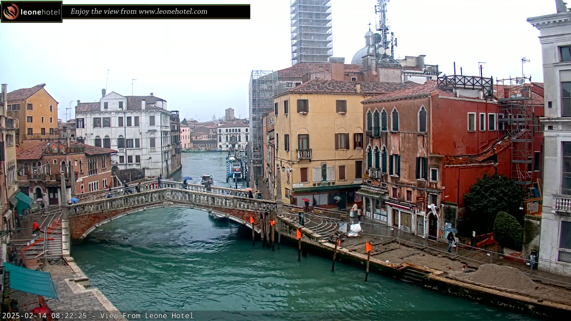 A view of a canal in Venice, Italy, on an overcast day, showing a stone bridge, colorful buildings, and scaffolding.