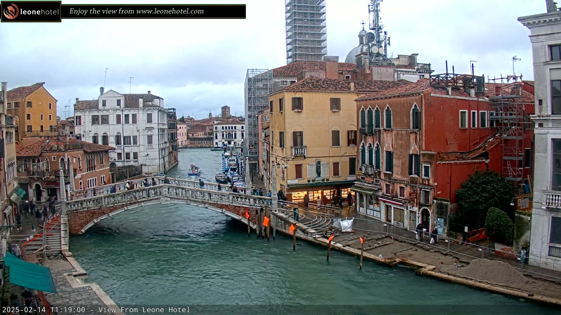 A stone bridge spans a canal in Venice, Italy, under an overcast sky; several boats are on the water, and buildings line the canal's edges.