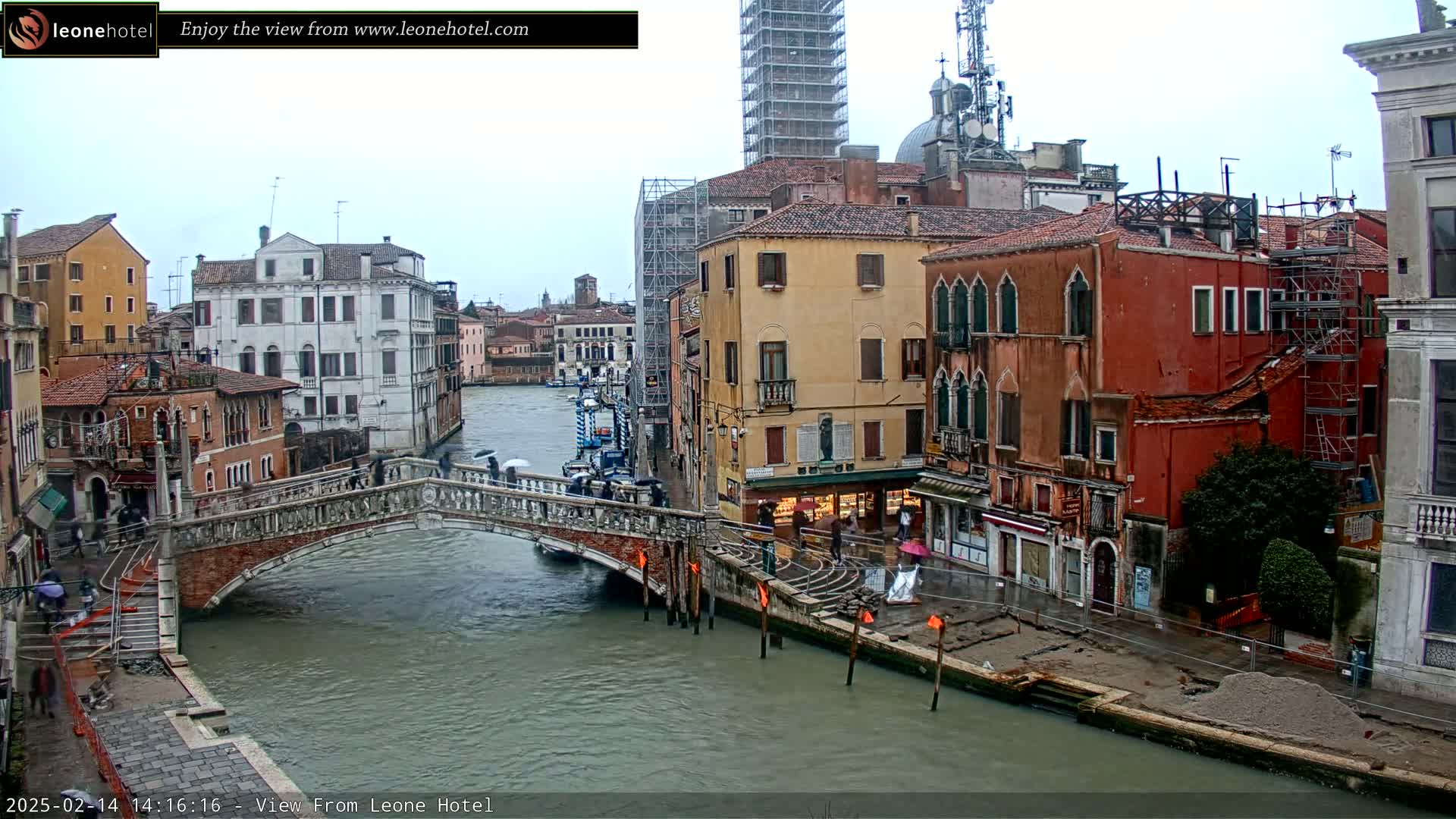 A view of a Venetian canal on a cloudy day, showing a stone bridge with people crossing it, colorful buildings lining the waterway, and scaffolding on some of the structures.