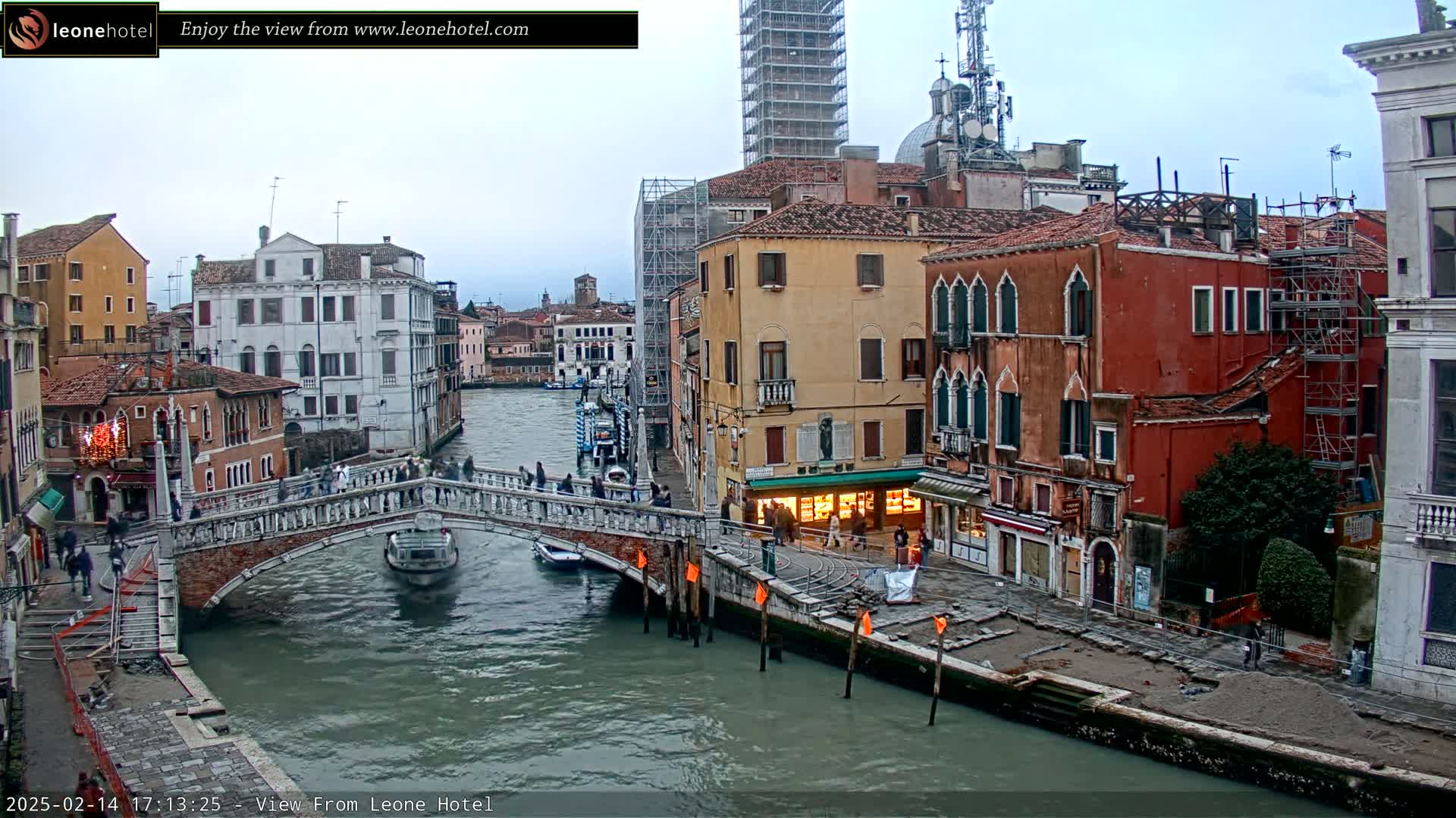 A canal in Venice, Italy, is shown under an overcast sky, with a stone bridge crossing the waterway and numerous buildings lining the banks.