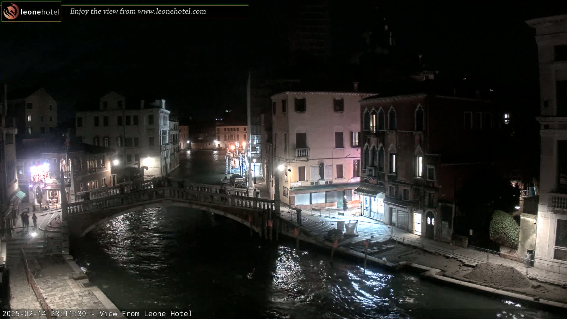 A nighttime view of a canal in Venice, showing a stone bridge and buildings lining the waterway under a dark sky.