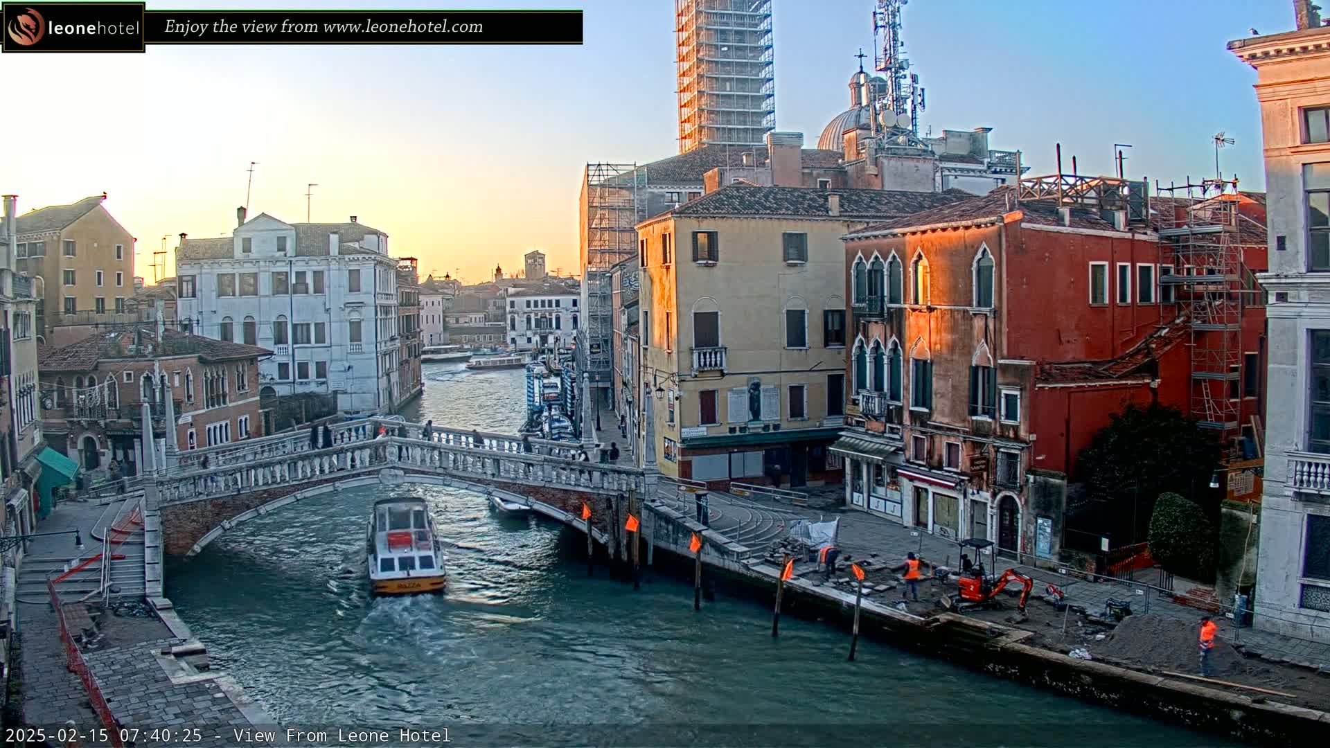 A canal in Venice, Italy, at sunrise, shows several buildings, a bridge, a small boat, and construction workers along the canal bank.