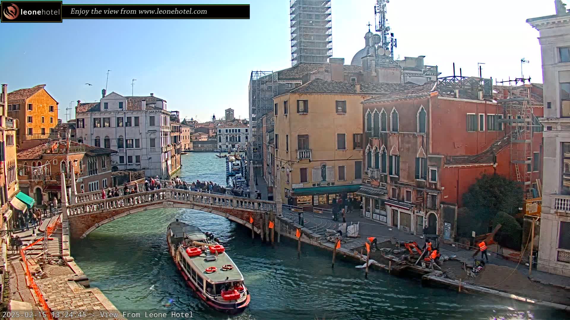 A sunny day in Venice shows a canal with a bridge crowded with people, boats, and buildings along the waterway, with construction work visible near the canal's edge.