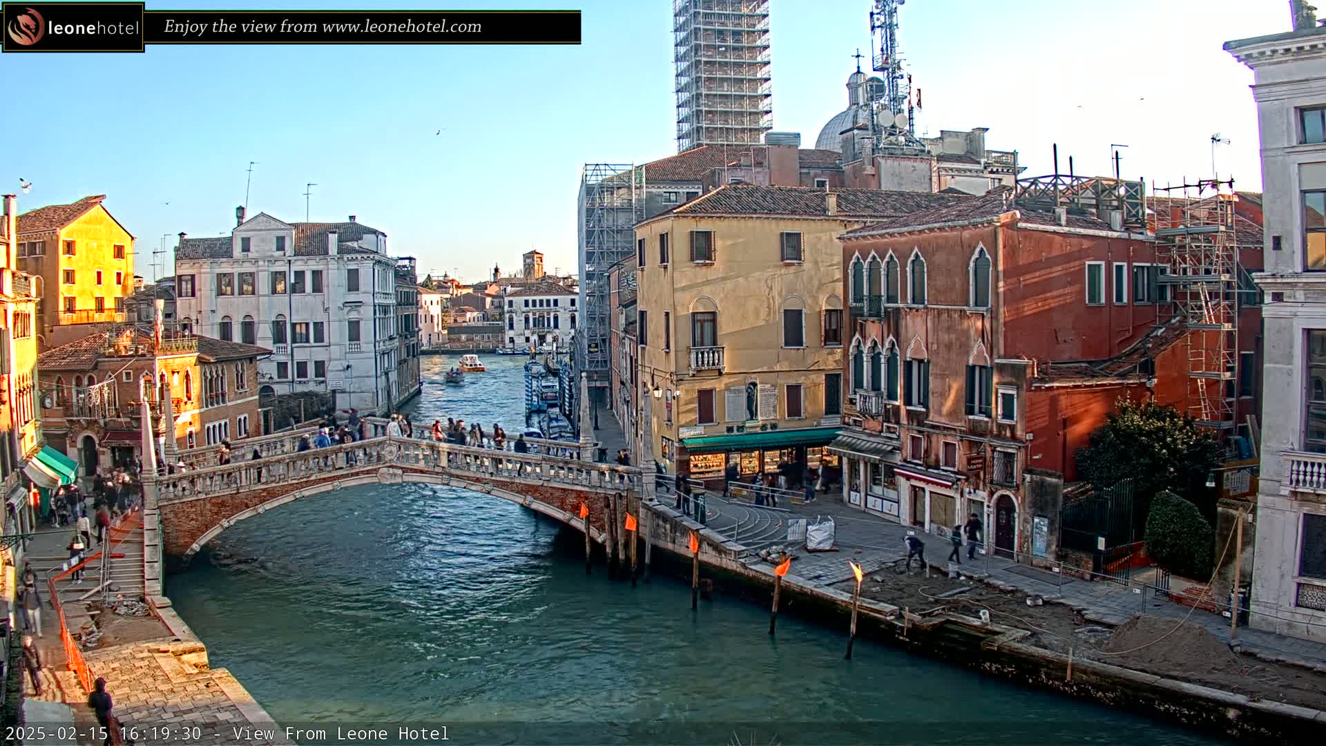 A stone bridge spans a canal in Venice, Italy, under a clear blue sky, with colorful buildings lining the waterway and pedestrians crossing the bridge.