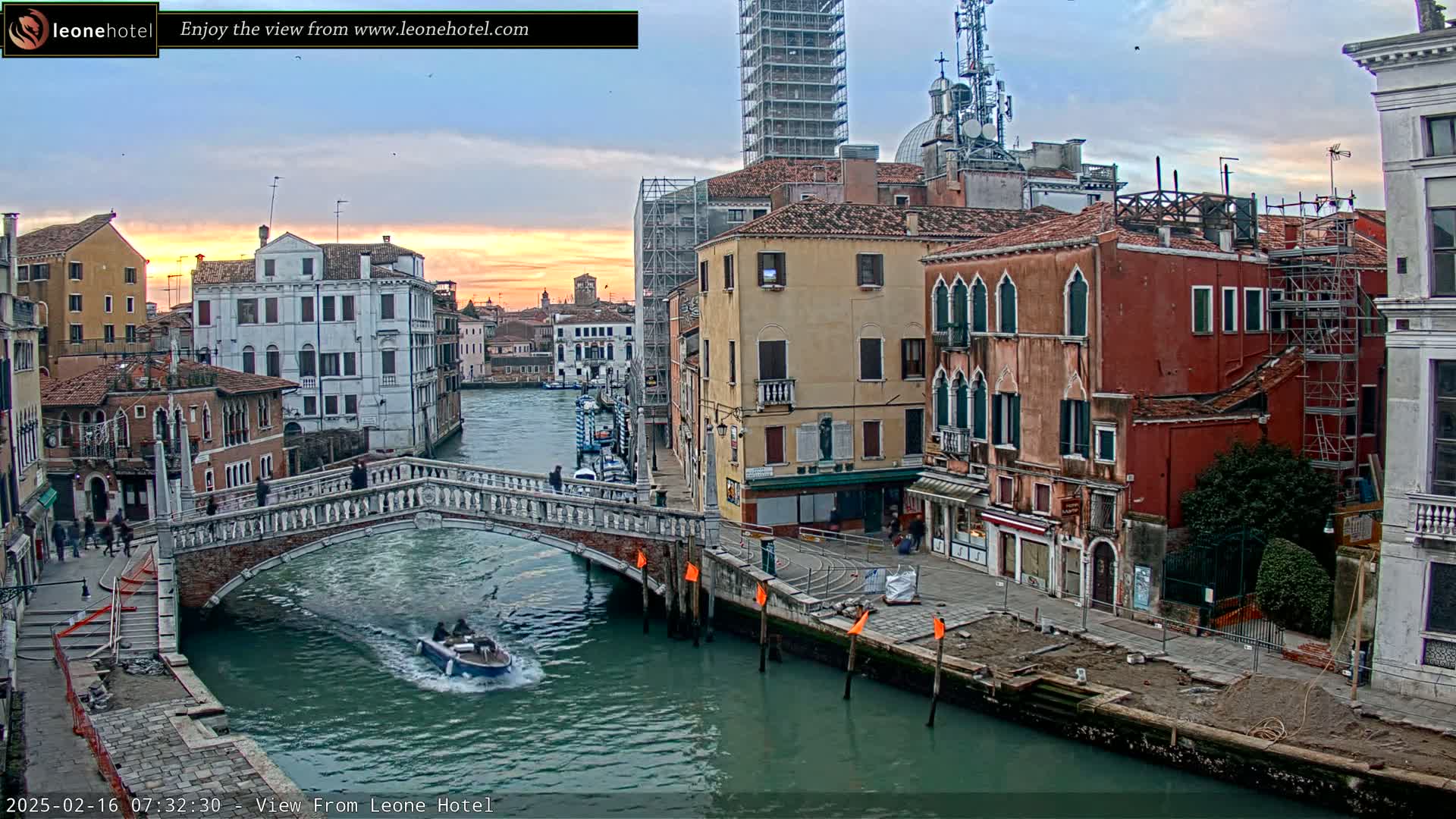 A Venetian canal at sunset, with a stone bridge, boats, and colorful buildings lining the waterway, under a partly cloudy sky.