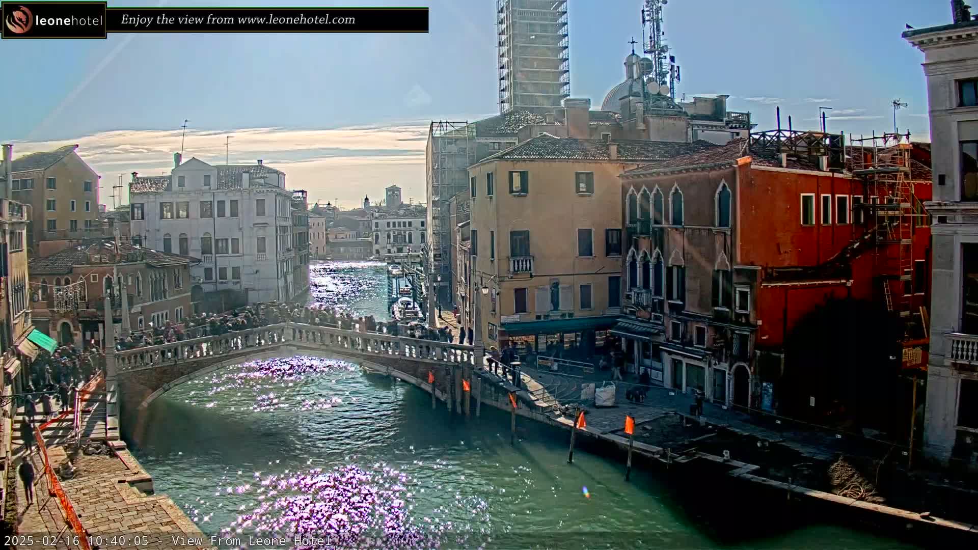 A sunny day in Venice shows a canal with a bridge crowded with people, and buildings lining the waterway, some under construction.