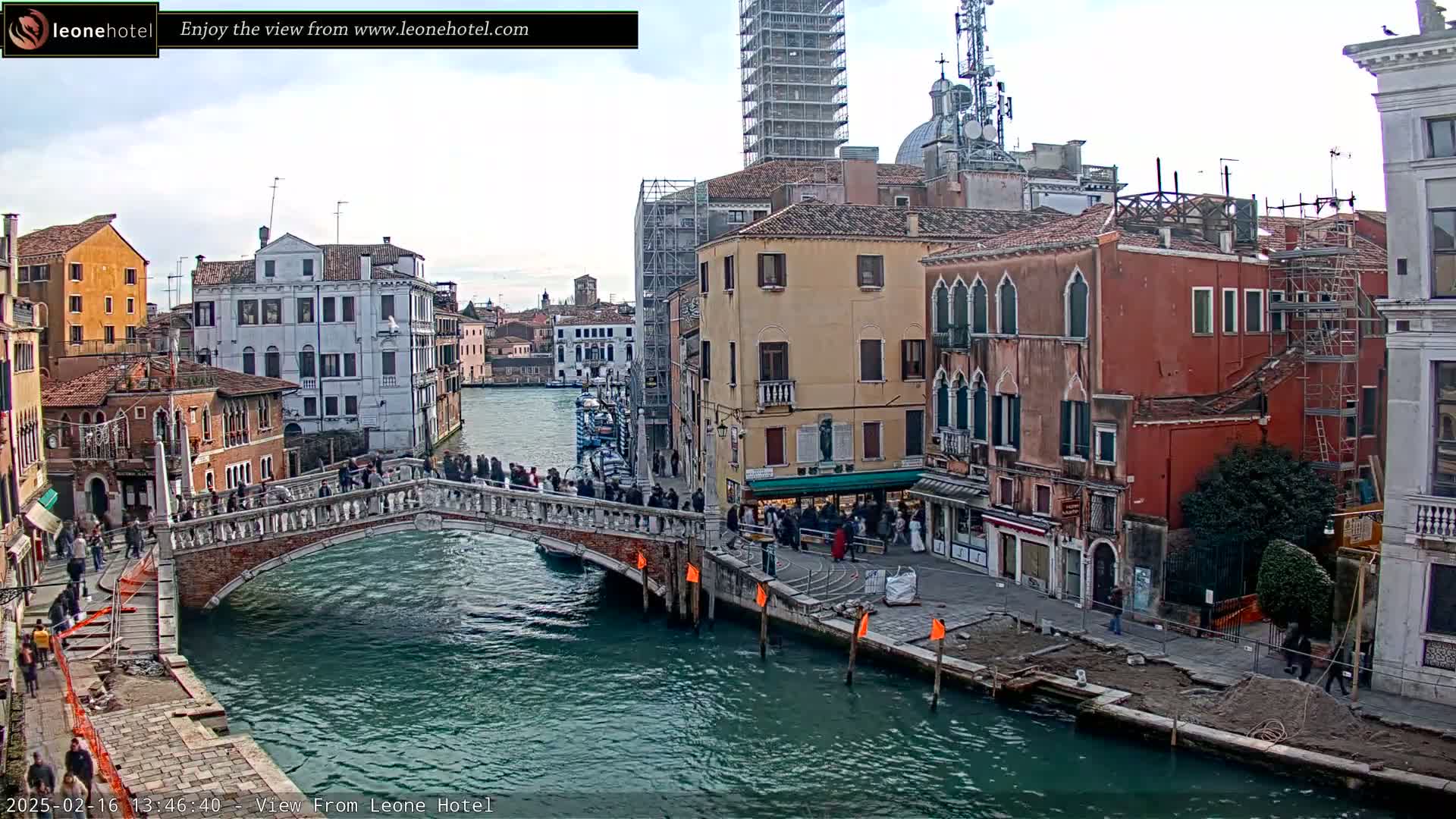 A stone bridge over a canal in Venice, Italy, is crowded with pedestrians under a cloudy sky.