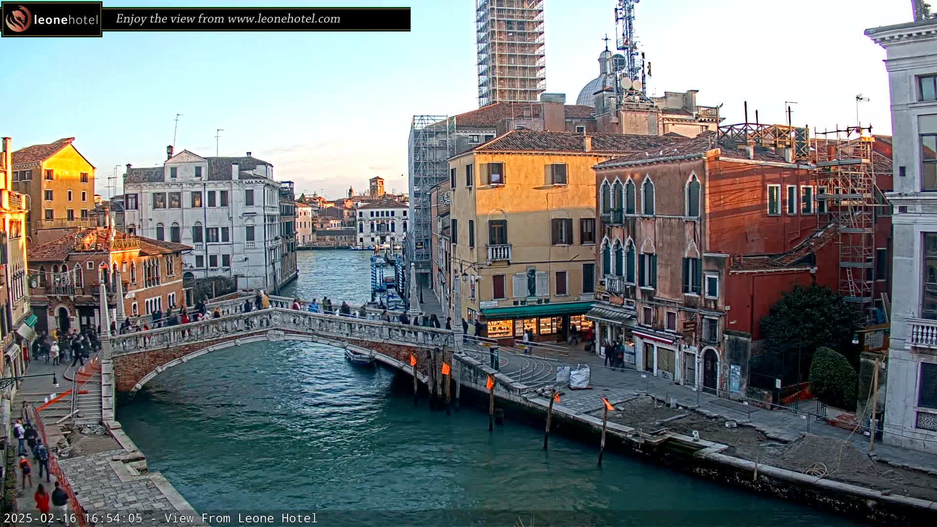 A stone bridge crosses a canal in Venice, Italy, under a clear, twilight sky, with pedestrians on the bridge and buildings lining the waterway.