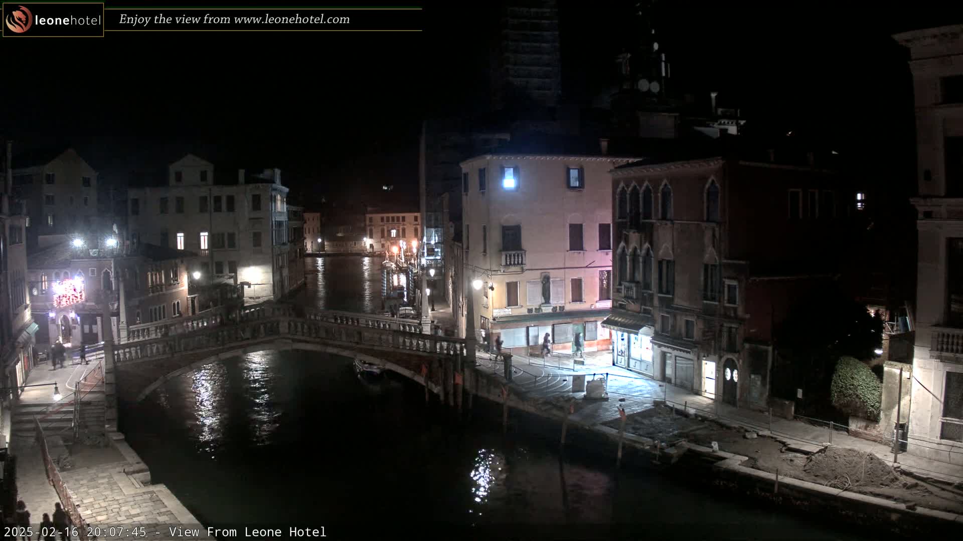 A nighttime view of a canal in Venice, Italy, shows a stone bridge, buildings lining the canal, and calm water reflecting the lights.