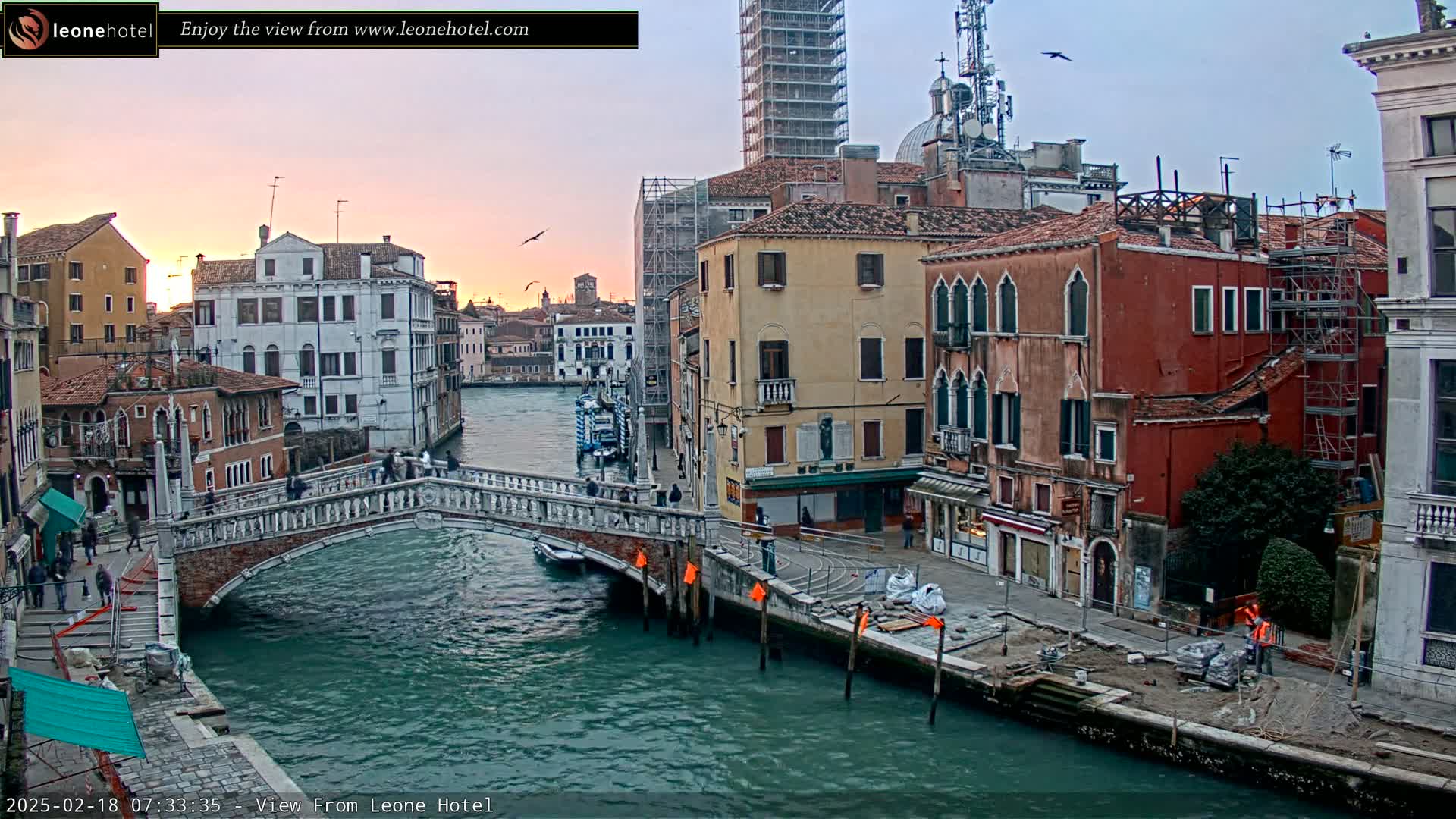 A canal in Venice at sunset, showing a stone bridge, several buildings, and construction along the waterway.