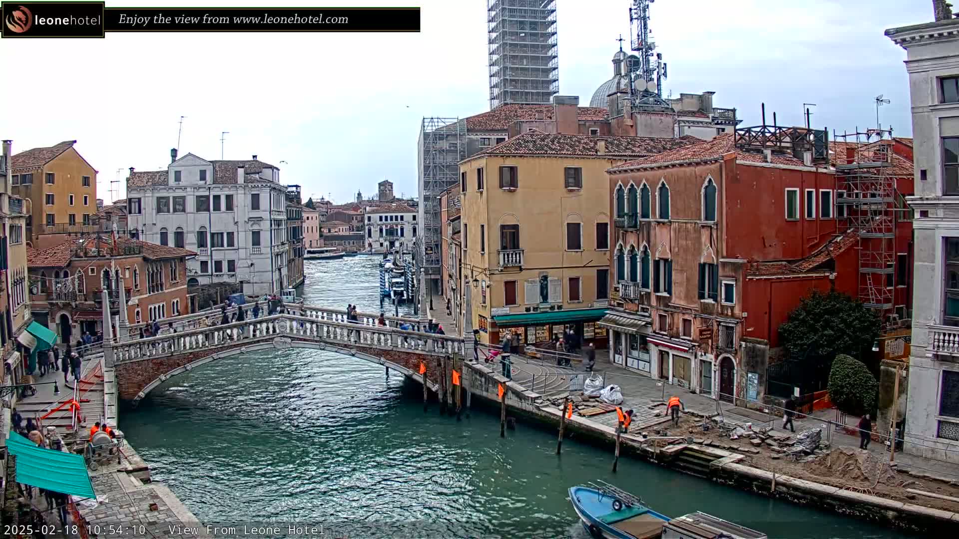 A bridge spans a canal in Venice, Italy, under an overcast sky, with colorful buildings lining the waterway and construction visible along the canal bank.