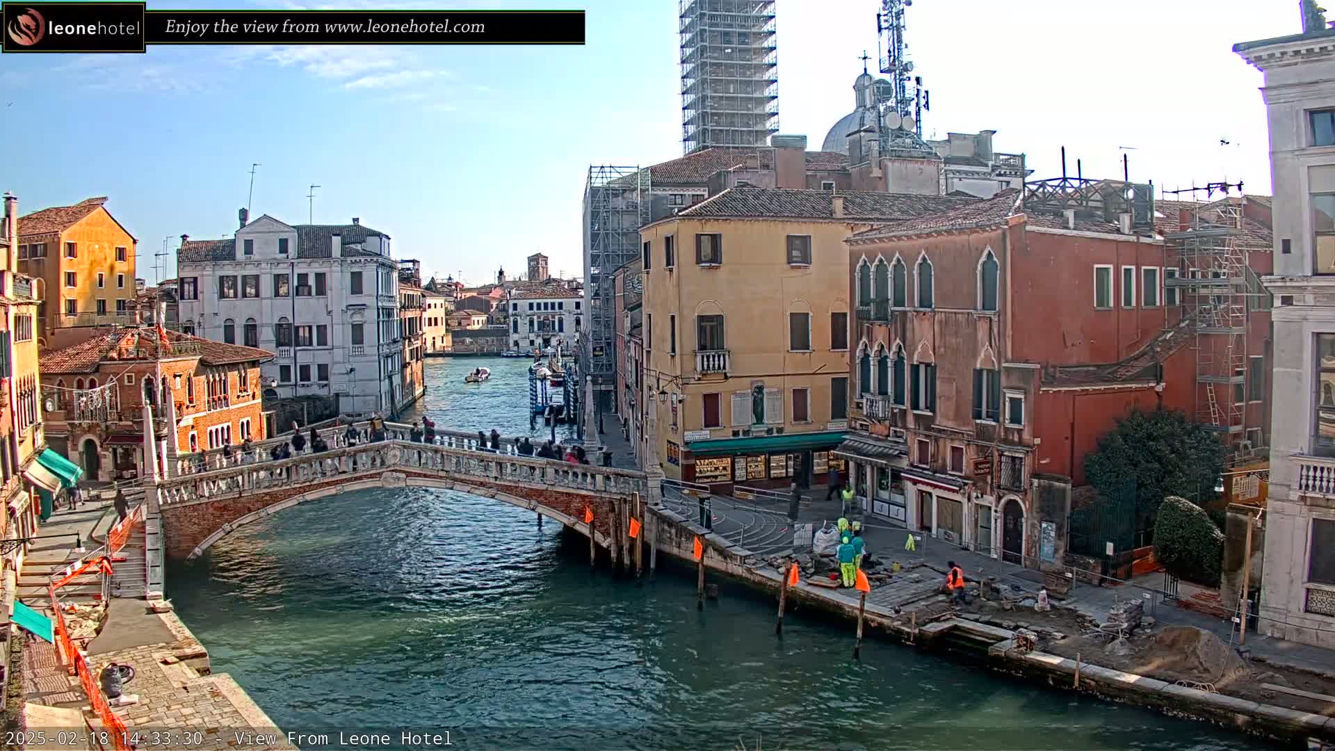 A sunny day in Venice shows a canal with a stone bridge, colorful buildings lining the waterway, and some construction work taking place along the canal bank.