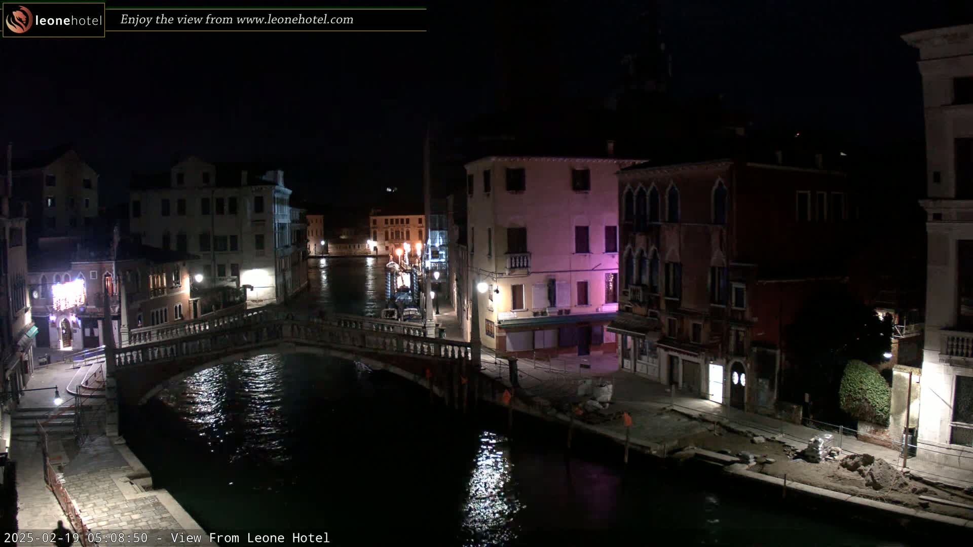 A nighttime view of a canal in Venice, Italy, shows a bridge and buildings lining the waterway under a dark sky.