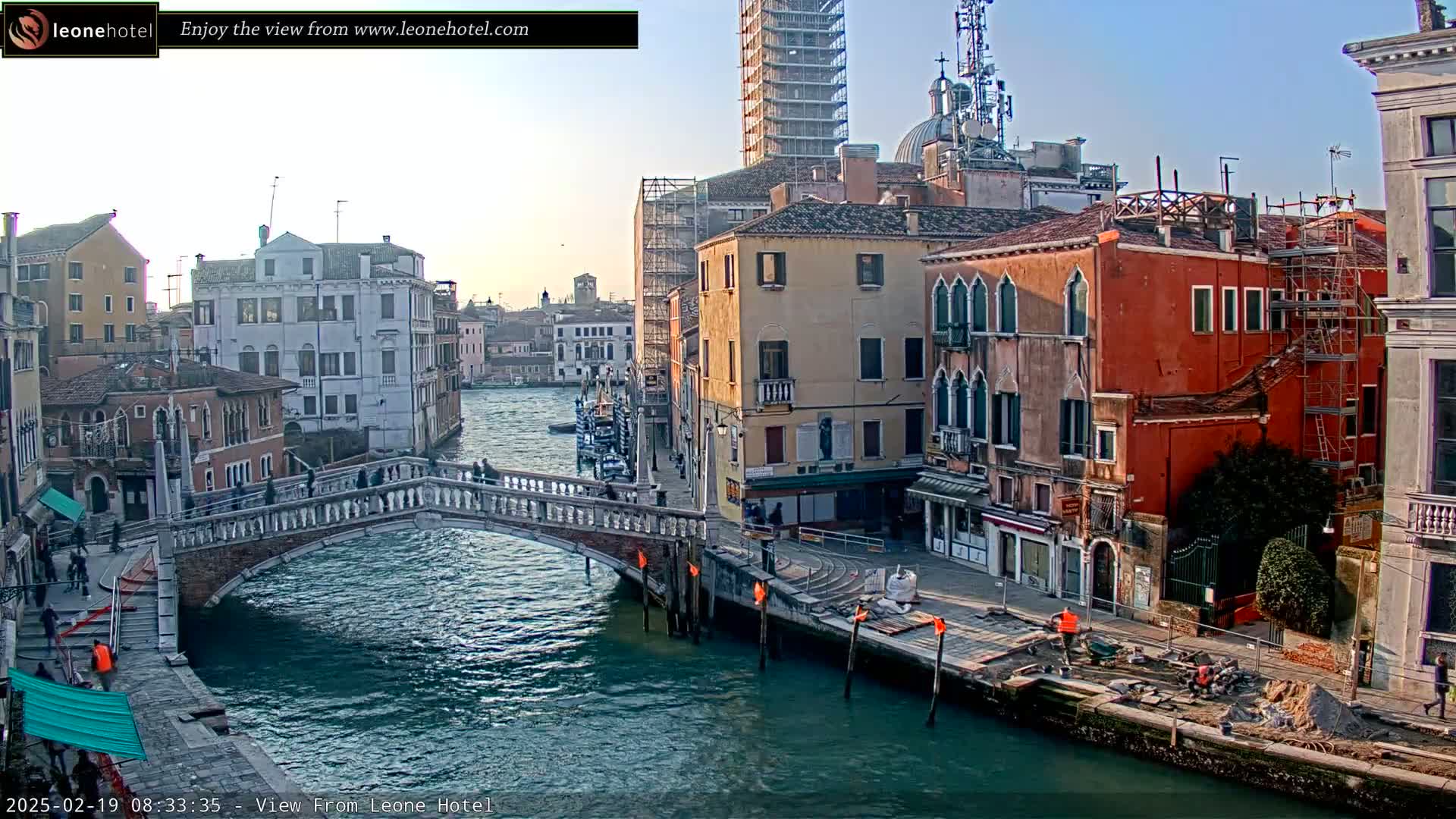 A Venetian canal scene under a clear, bright sky shows a bridge with pedestrians, colorful buildings lining the waterway, and some construction activity along the canal bank.