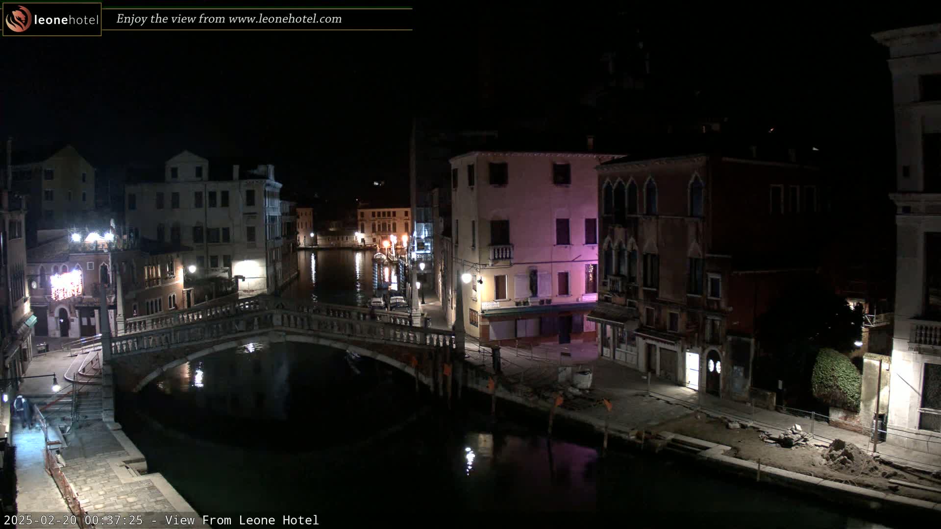 A nighttime view of a canal in Venice, Italy, shows a stone bridge over calm water, with buildings lining the canals on both sides under a clear, dark sky.