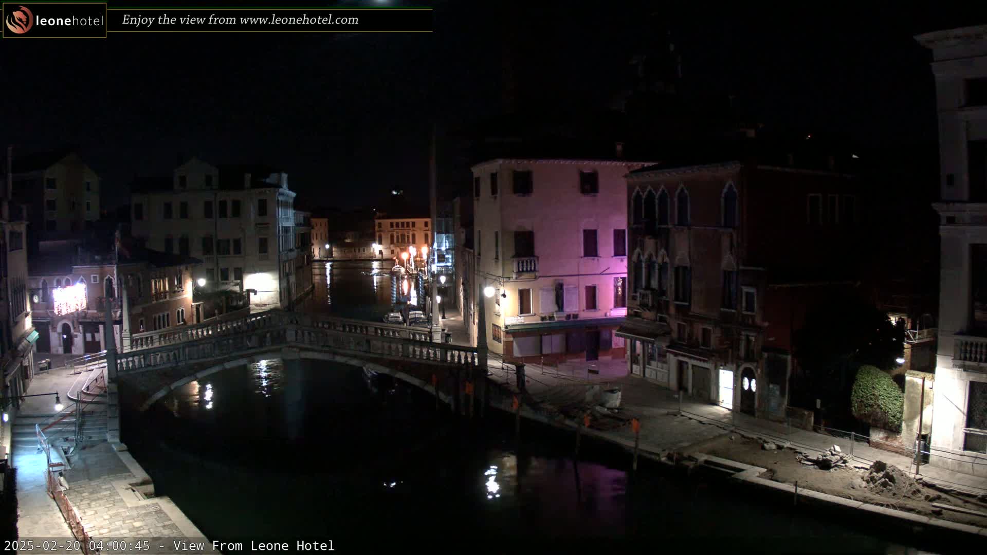 A nighttime view of a canal in Venice, Italy, shows a stone bridge over the water, with buildings lining both sides of the canal, under a dark sky.