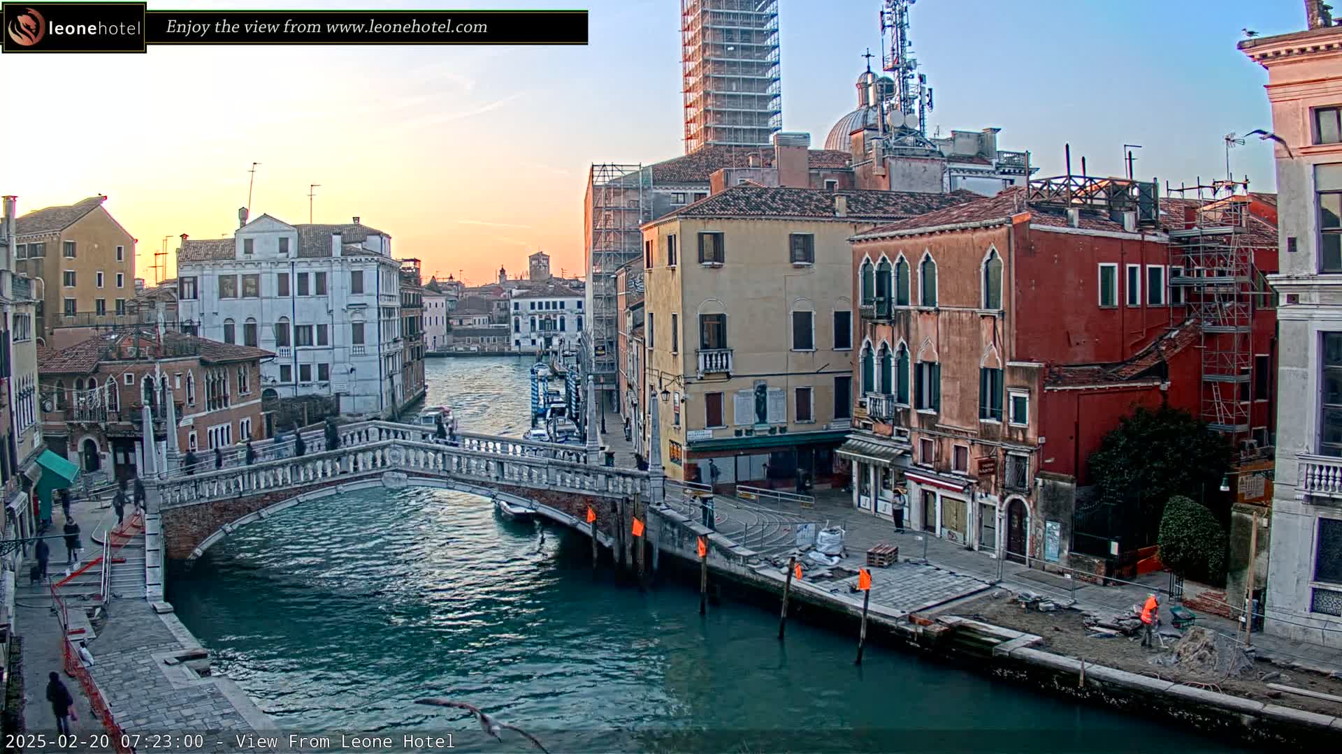 A stone bridge spans a calm canal in Venice at sunset, with colorful buildings lining the waterway and construction workers visible along the canal's edge.