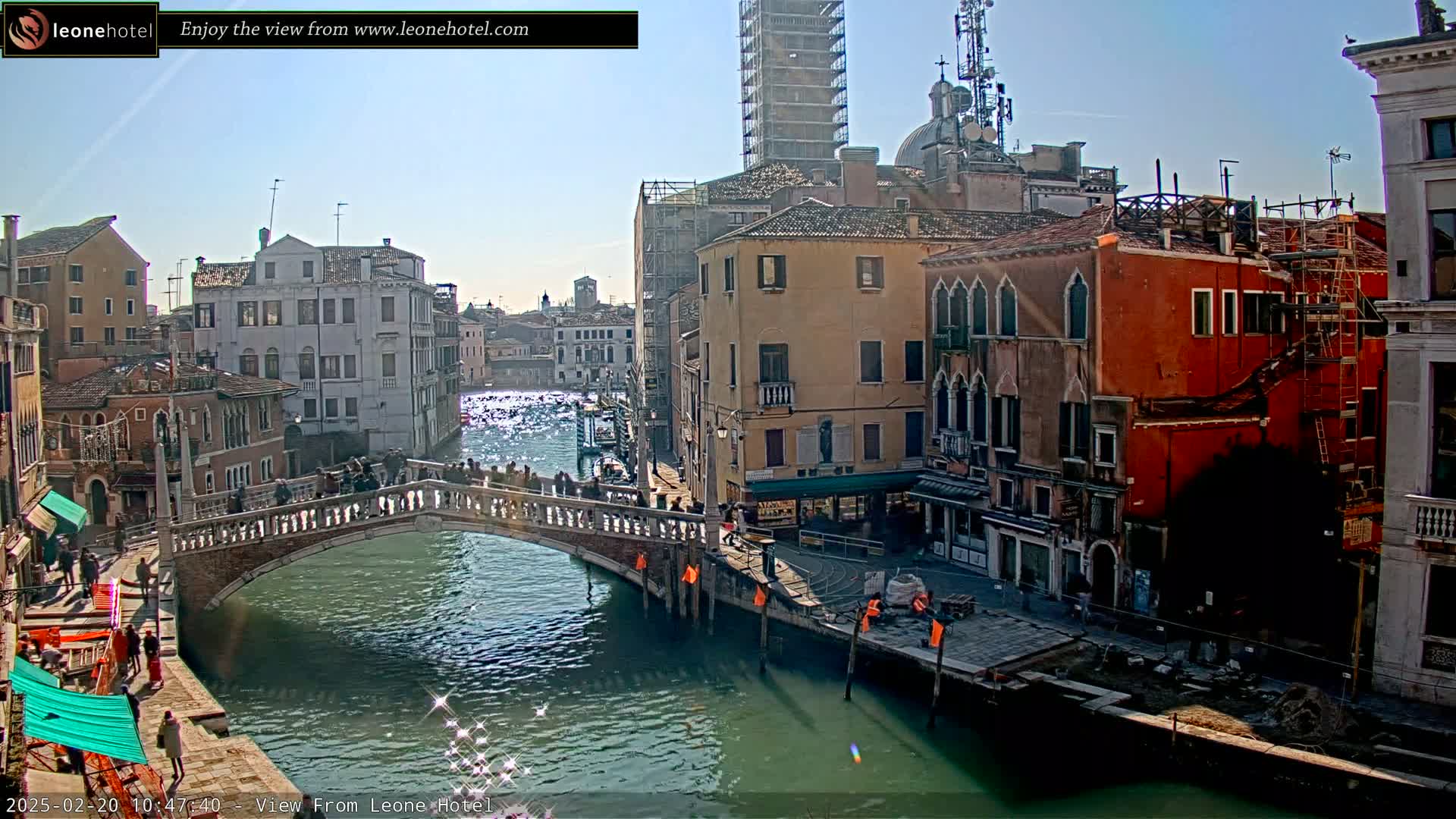 A sunny day in Venice shows a canal with a stone bridge, several buildings along the waterway, and some people walking on the bridge and sidewalks.