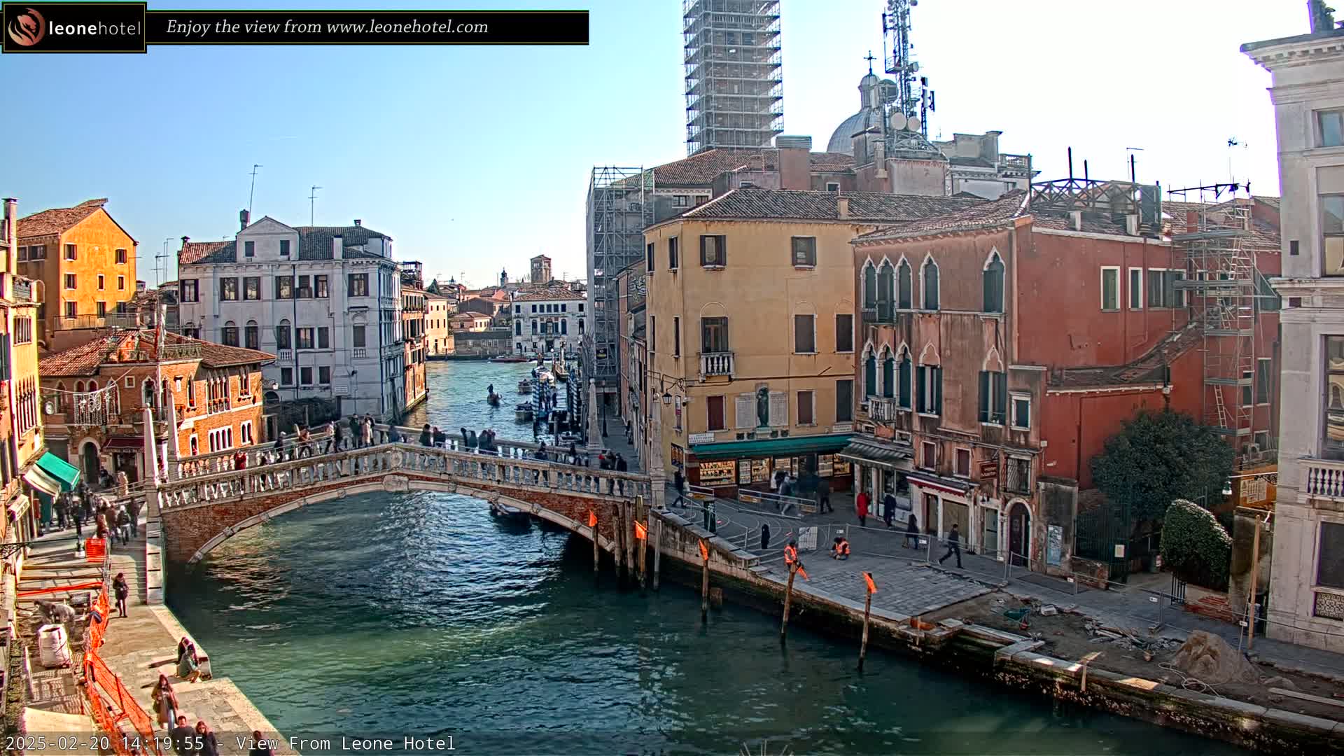 A sunny day in Venice shows a canal with a stone bridge, pedestrians walking on it and along the canal-side, and colorful buildings lining the waterway, some under construction.