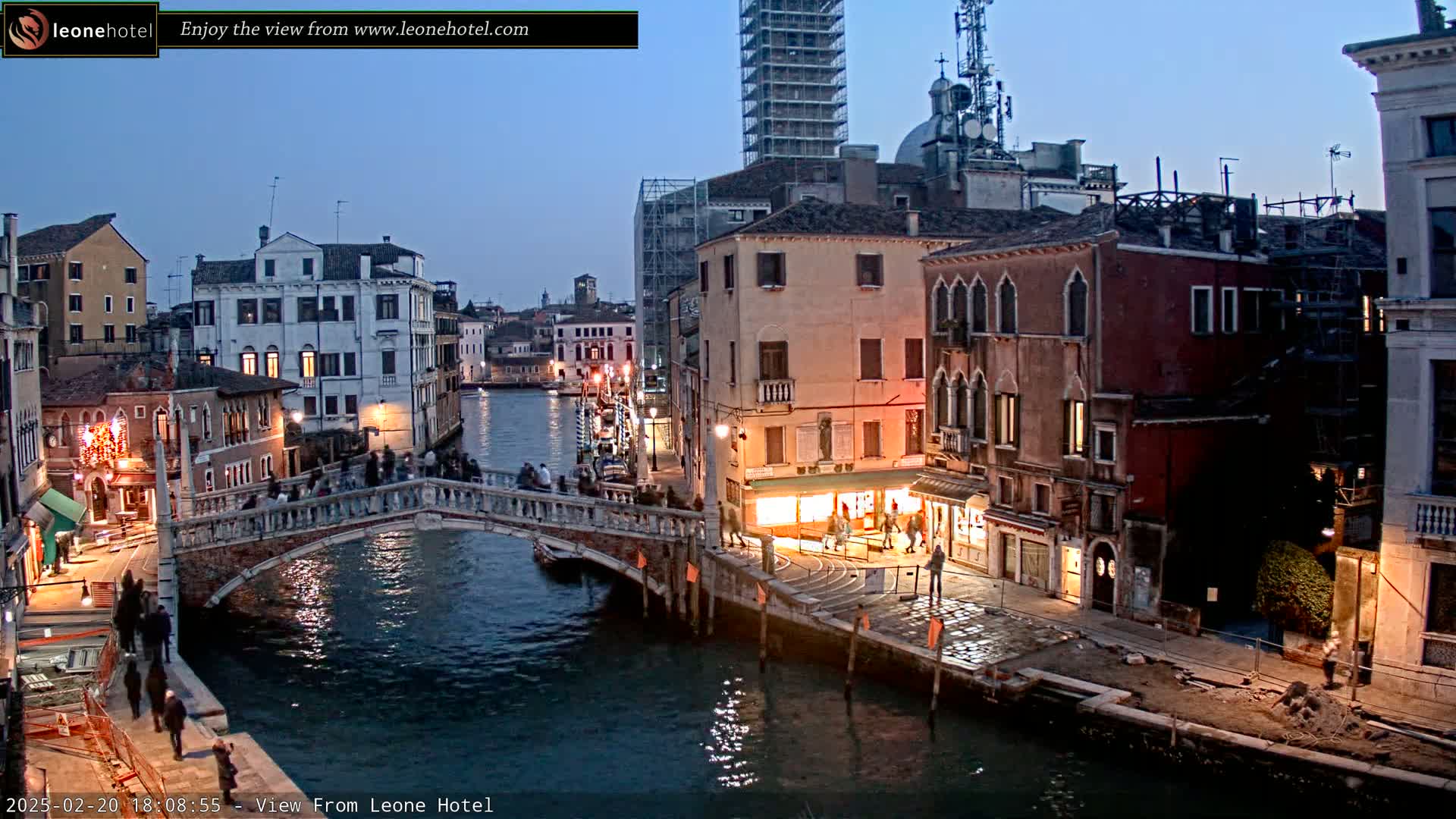 A bridge over a canal in Venice at twilight, with illuminated buildings lining the waterway and people walking on the bridge and the canal-side walkways.