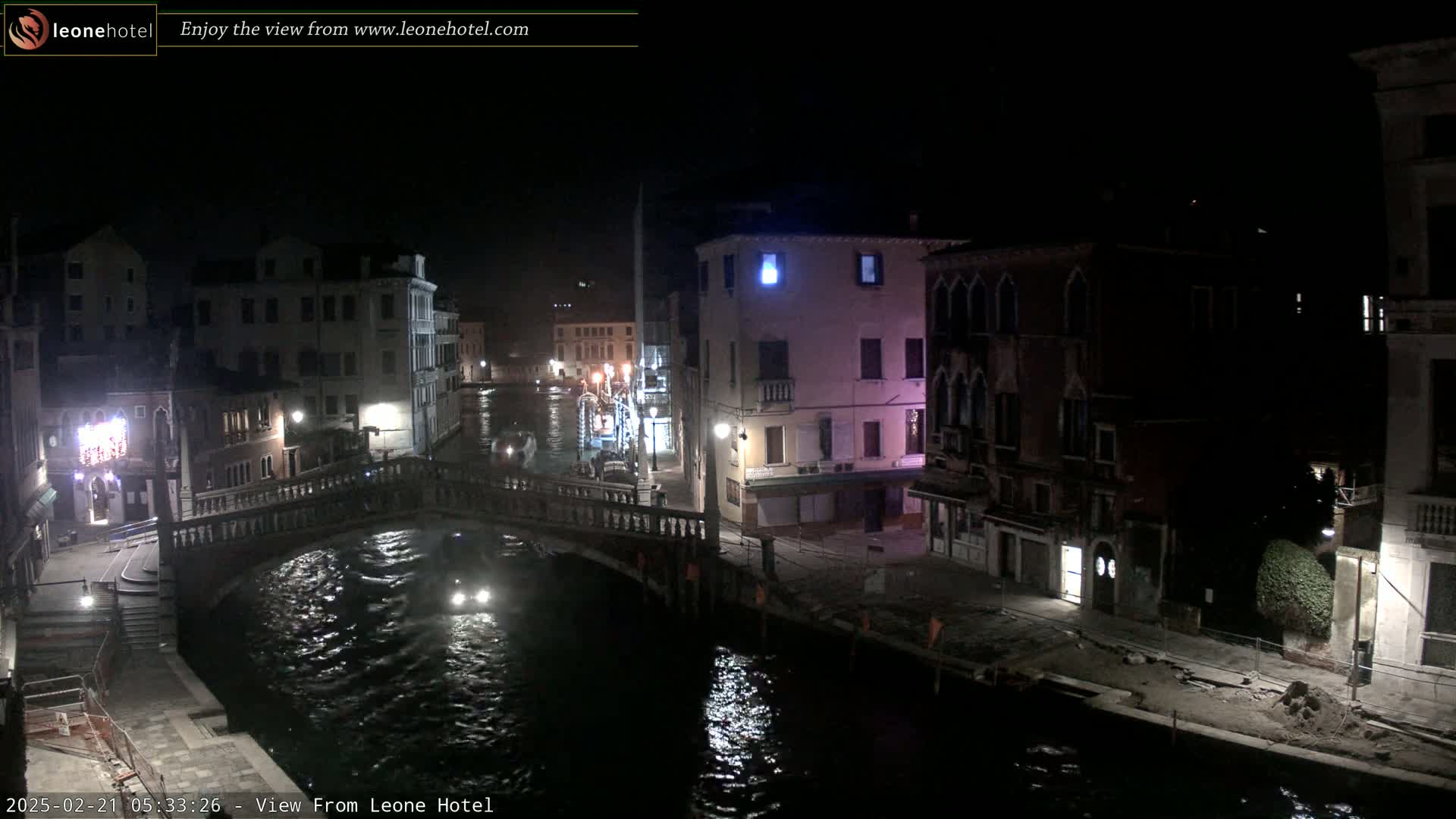 A nighttime view of a canal in Venice, showing buildings lining the waterway and a bridge, with a boat on the water.