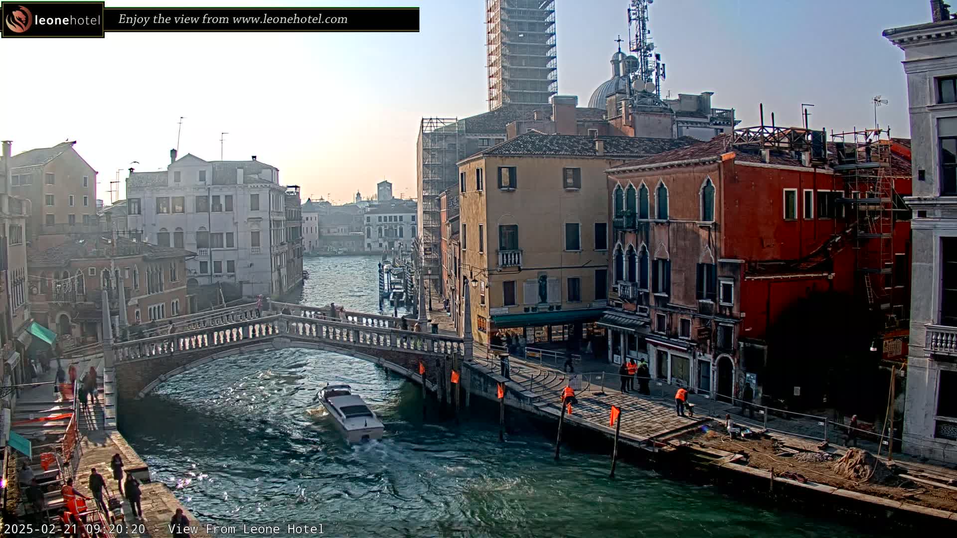 A Venetian canal scene under a bright, sunny sky shows a bridge, several buildings, a motorboat, and people walking along the canal's edge.