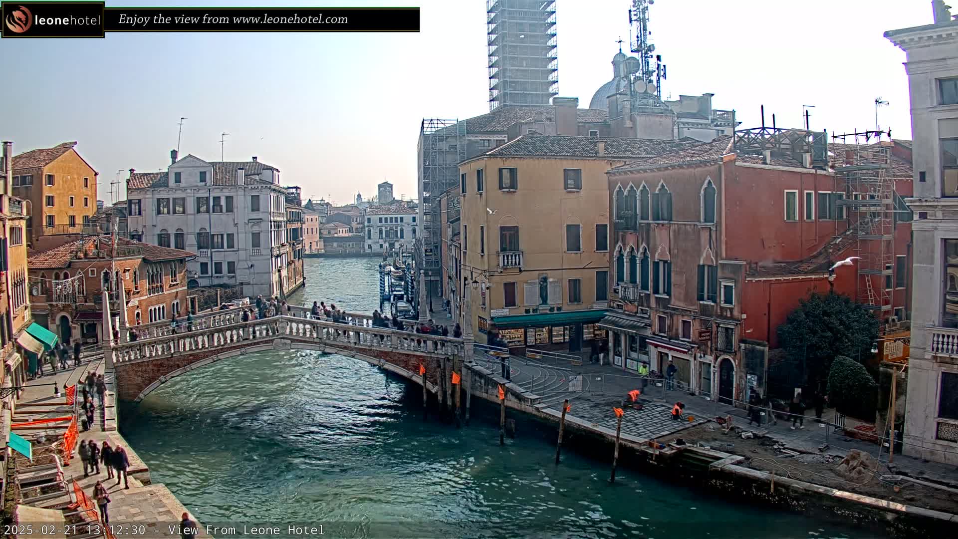 A bridge over a canal in Venice, Italy, on a sunny day, with numerous buildings lining the waterway, some under construction.