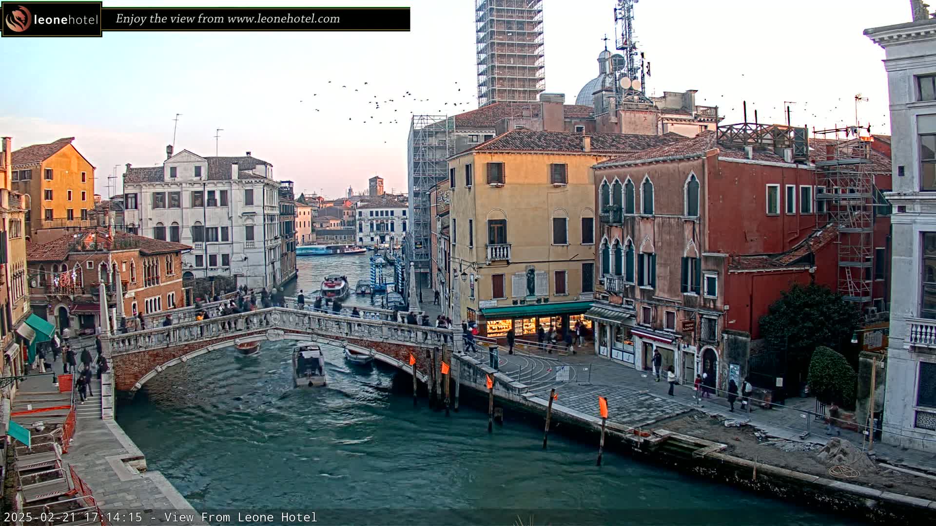 A bridge over a canal in Venice, Italy, is surrounded by colorful buildings under a twilight sky with a flock of birds in flight.