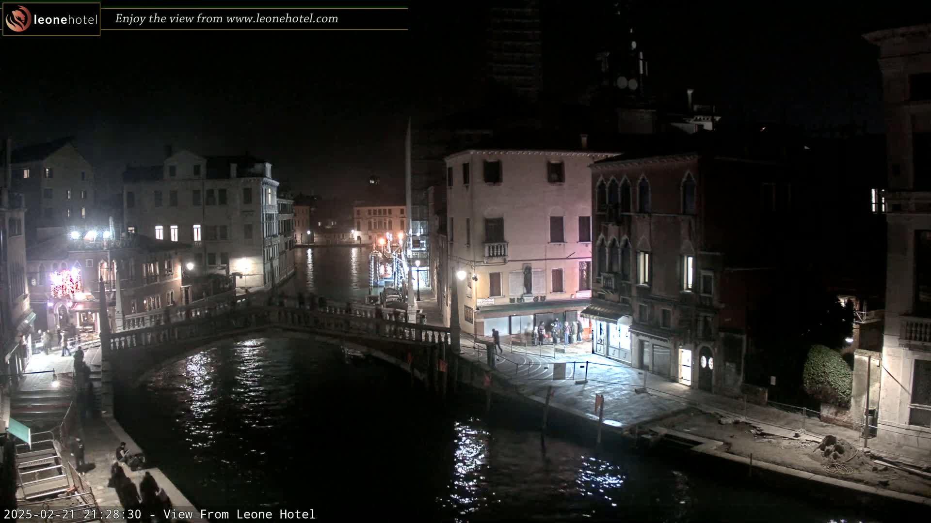 A nighttime view of a Venetian canal, showing bridges, buildings, and a few people along the water's edge.