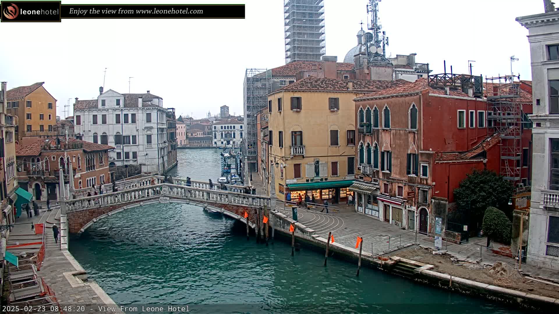 A view of a canal in Venice, Italy, on an overcast day, shows a stone bridge spanning the waterway between colorful buildings, some of which are under construction.