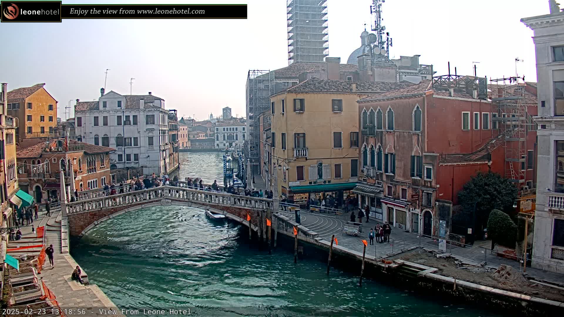 A canal in Venice, Italy, under a cloudy sky, is spanned by a bridge with people walking across it, and lined with historic buildings, some of which are under construction.