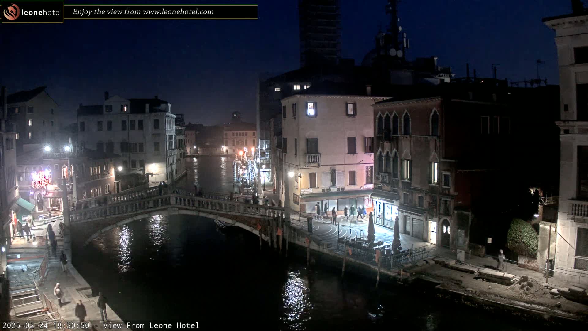 A nighttime view of a canal in Venice, Italy, shows a bridge, several buildings, and a few pedestrians.