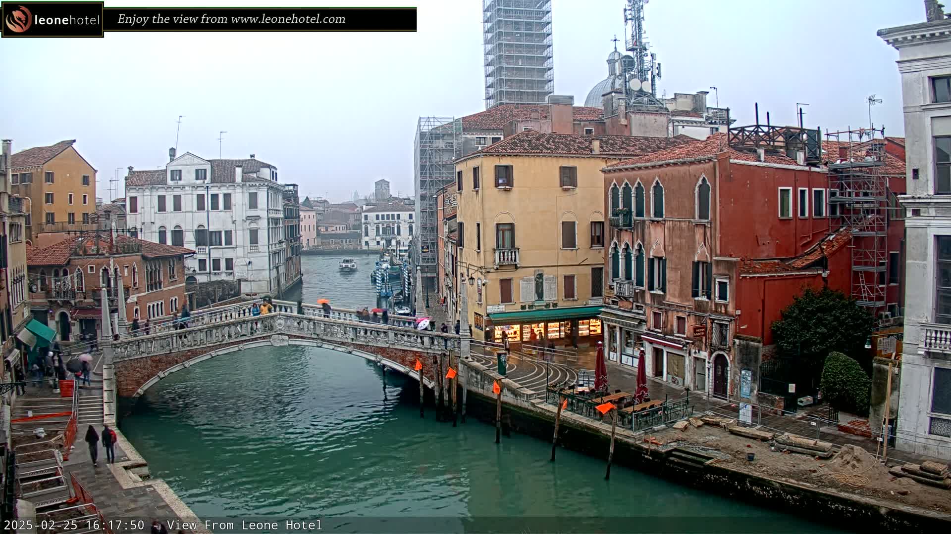 A stone bridge spans a canal in Venice, Italy, under an overcast sky, with colorful buildings lining the waterway and scaffolding visible on some structures.