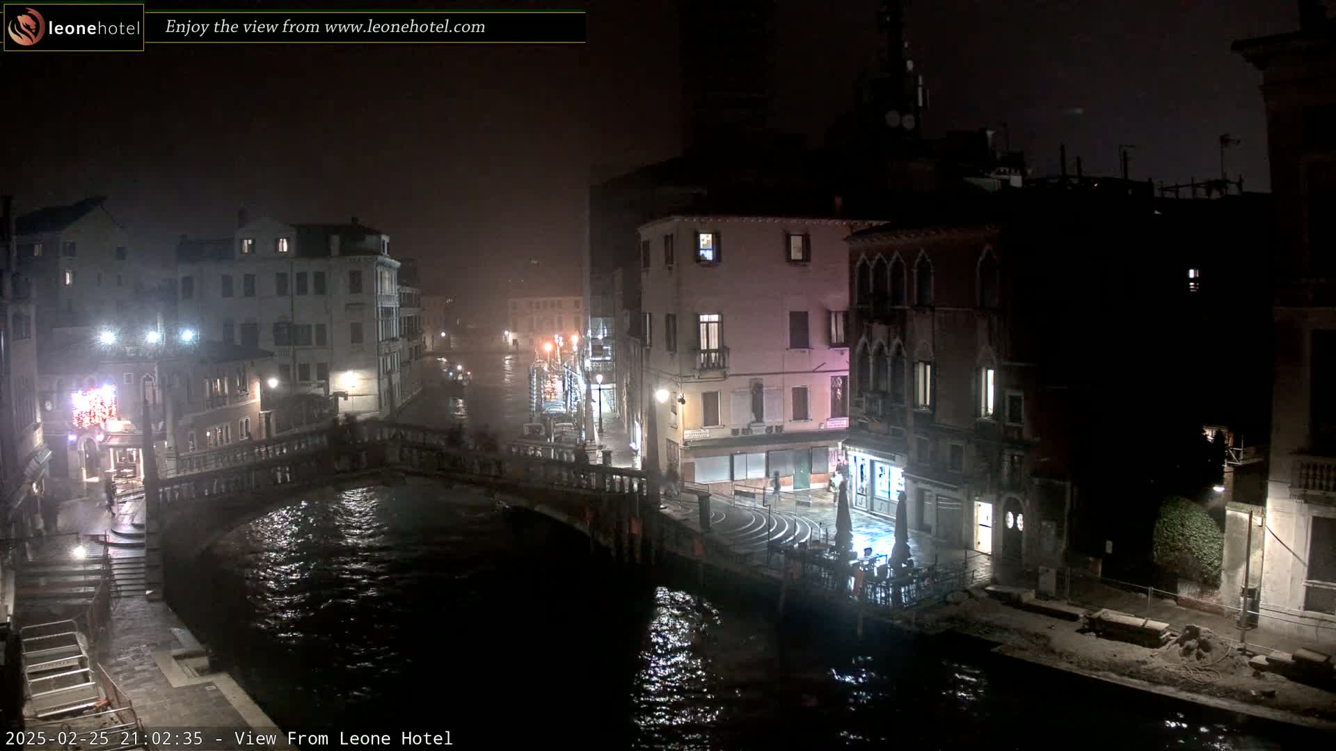 A nighttime view of a canal in Venice, Italy, shrouded in fog or mist, shows several buildings lining the waterway and a bridge spanning the canal.