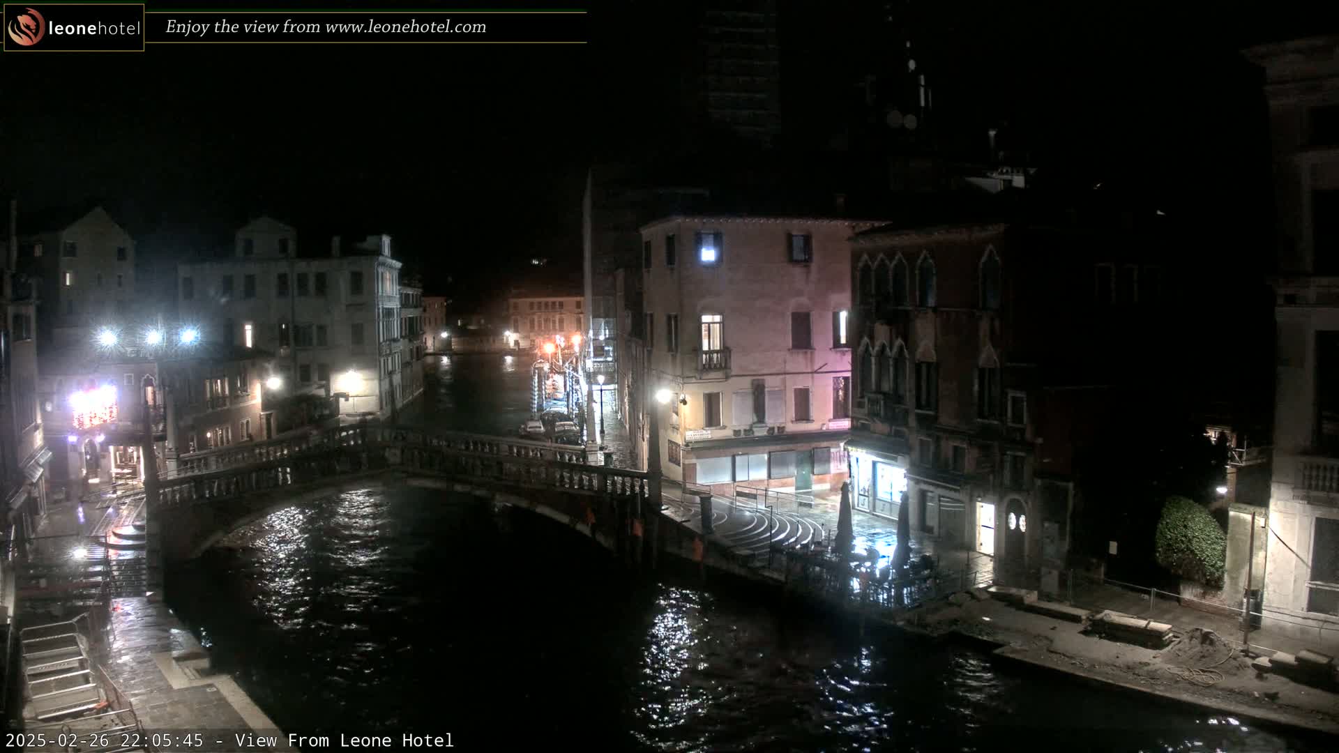 A nighttime view of a canal in Venice, showing several buildings and a bridge, with wet pavement suggesting rain or recent rain.