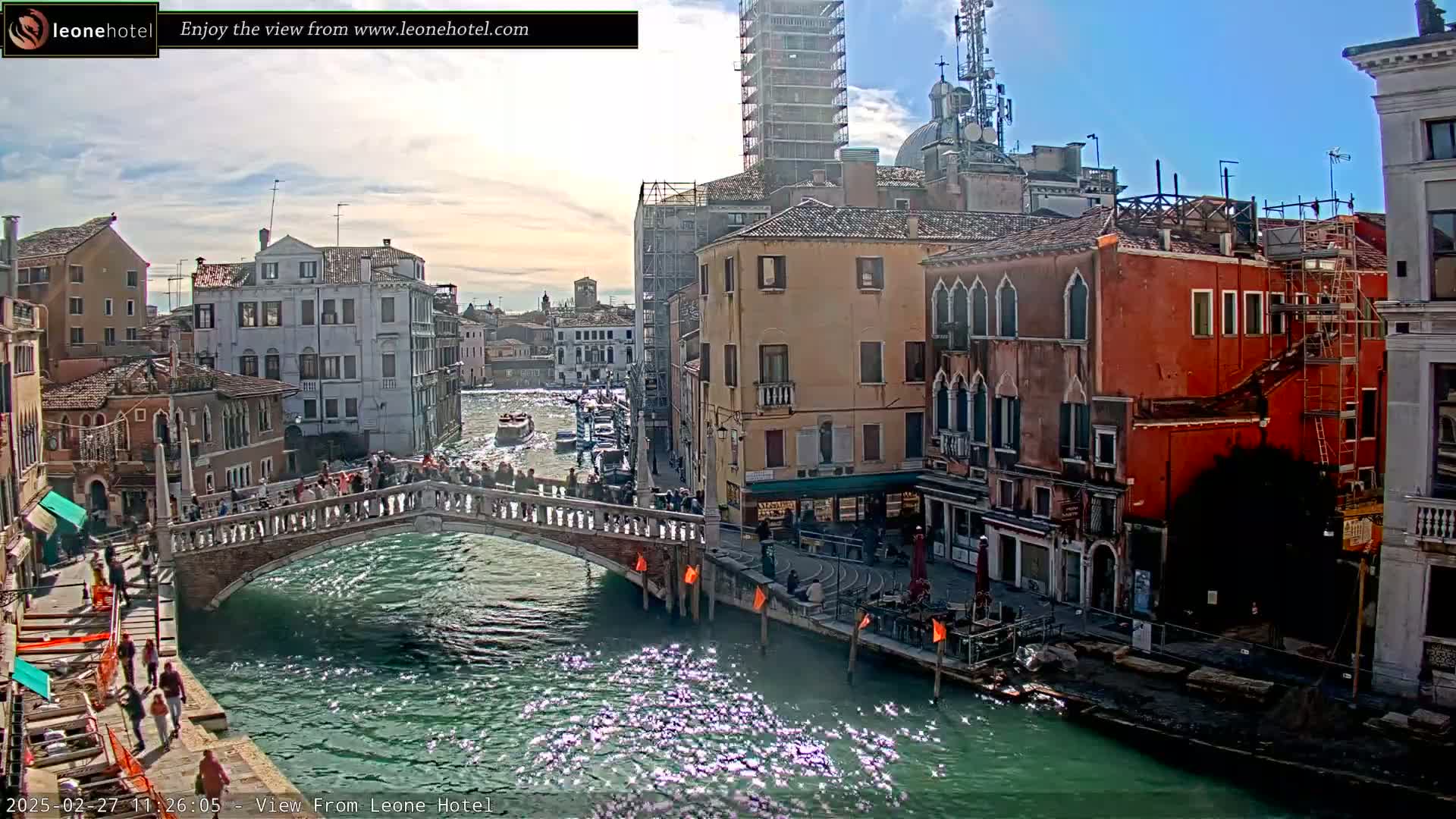 A sunny day in Venice shows a canal with a bridge crowded with people, boats on the water, and colorful buildings lining the waterway, some under construction.