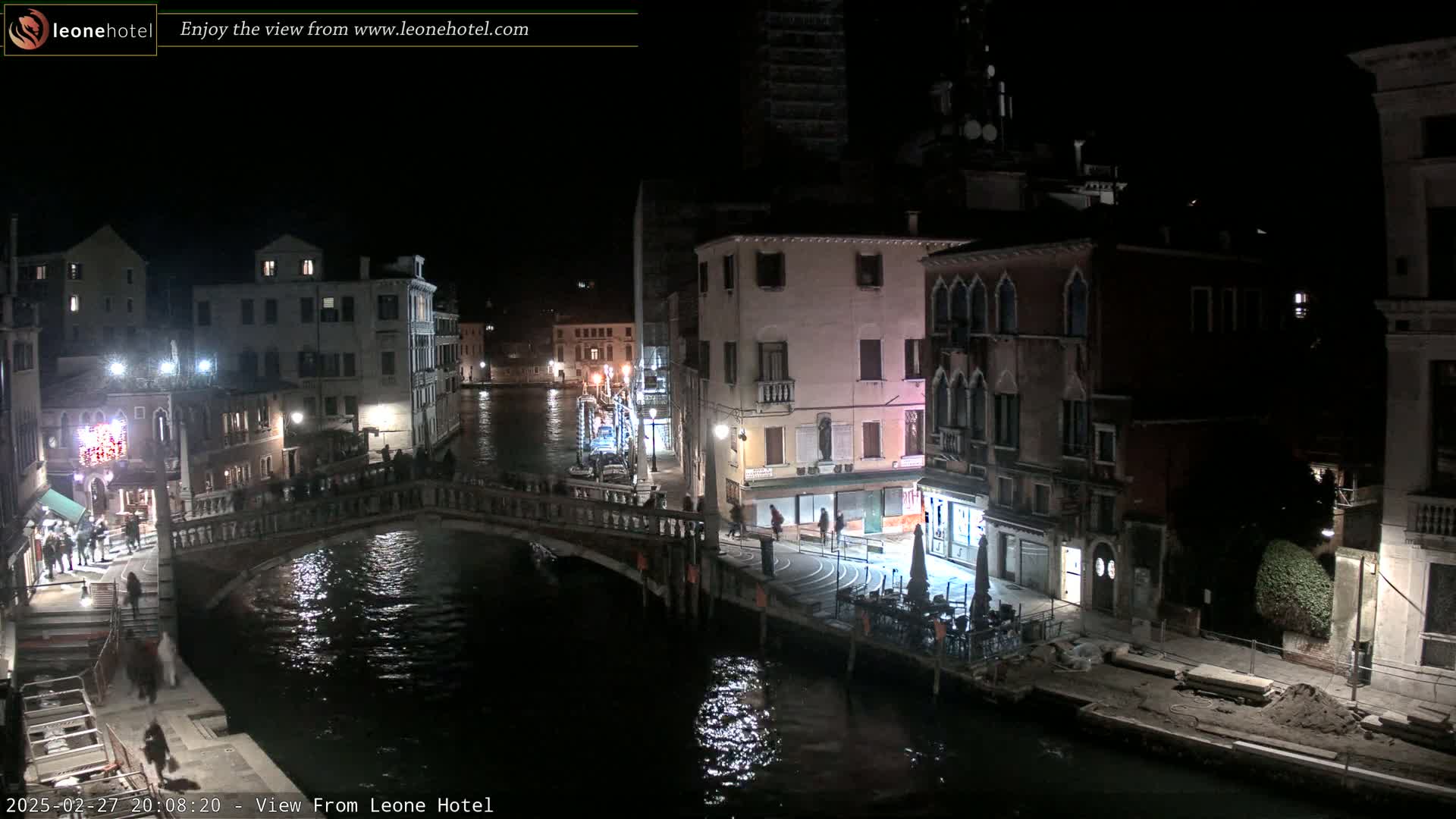 A nighttime view of a Venetian canal, showing several buildings lit up along the waterway, a bridge with people on it, and a calm canal reflecting the lights.