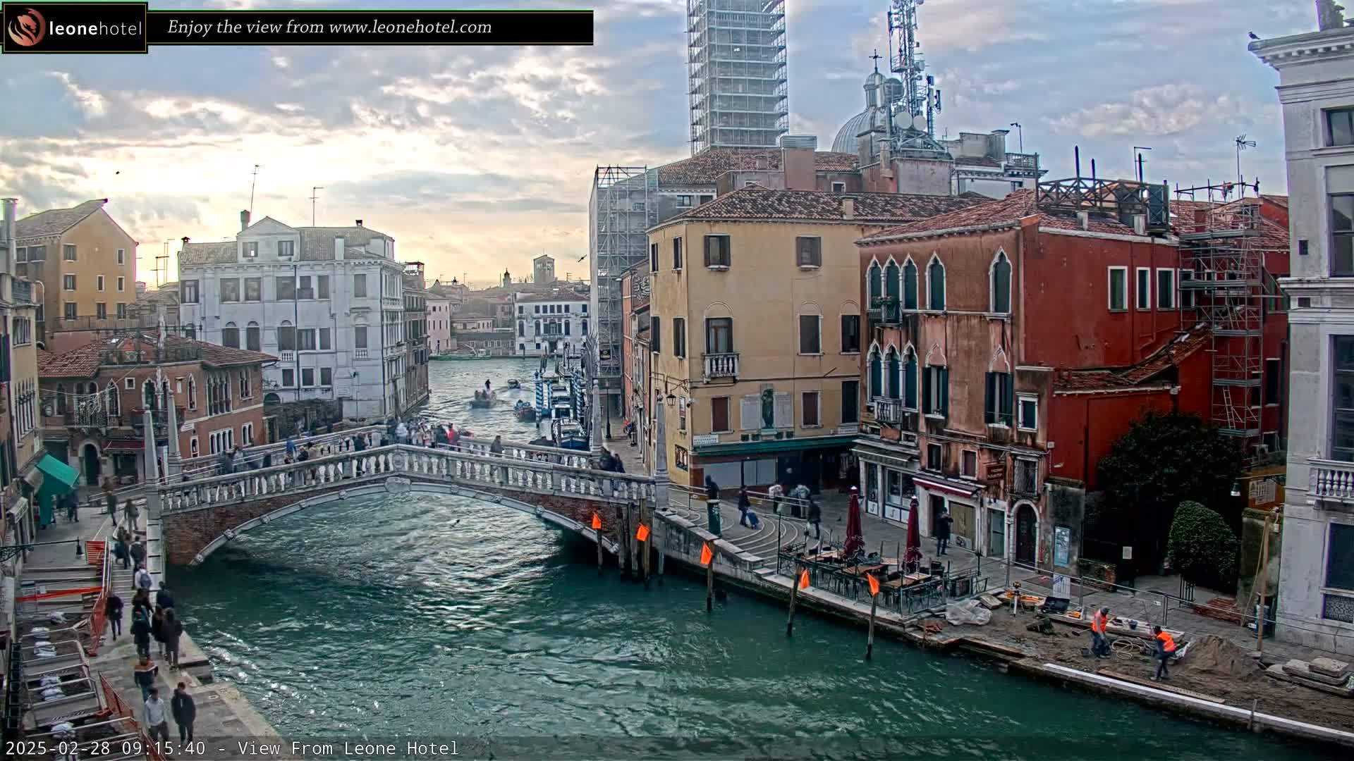 A canal in Venice, Italy, under a partly cloudy sky, shows a bridge with pedestrians crossing it, boats on the water, and several buildings along the waterway, with some construction work visible.