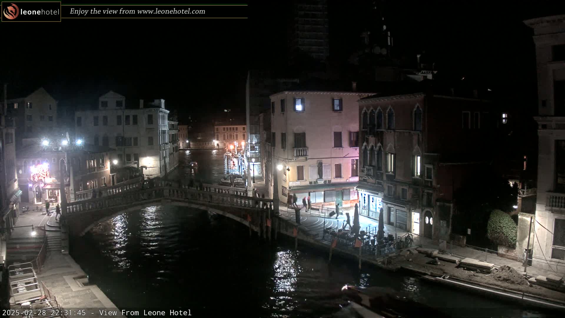 A nighttime view of a canal in Venice, Italy, shows a bridge, buildings lining the waterway, and a few people walking along the canal.