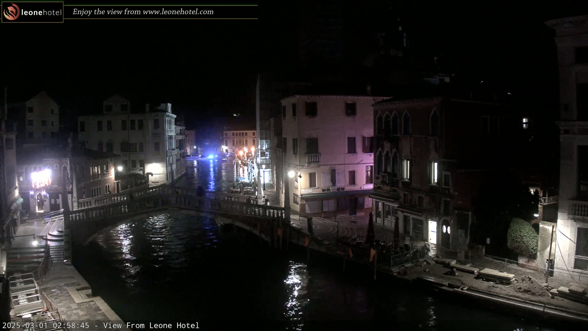 A nighttime view of a canal in Venice, Italy, shows several buildings along the waterway and a bridge spanning the canal.