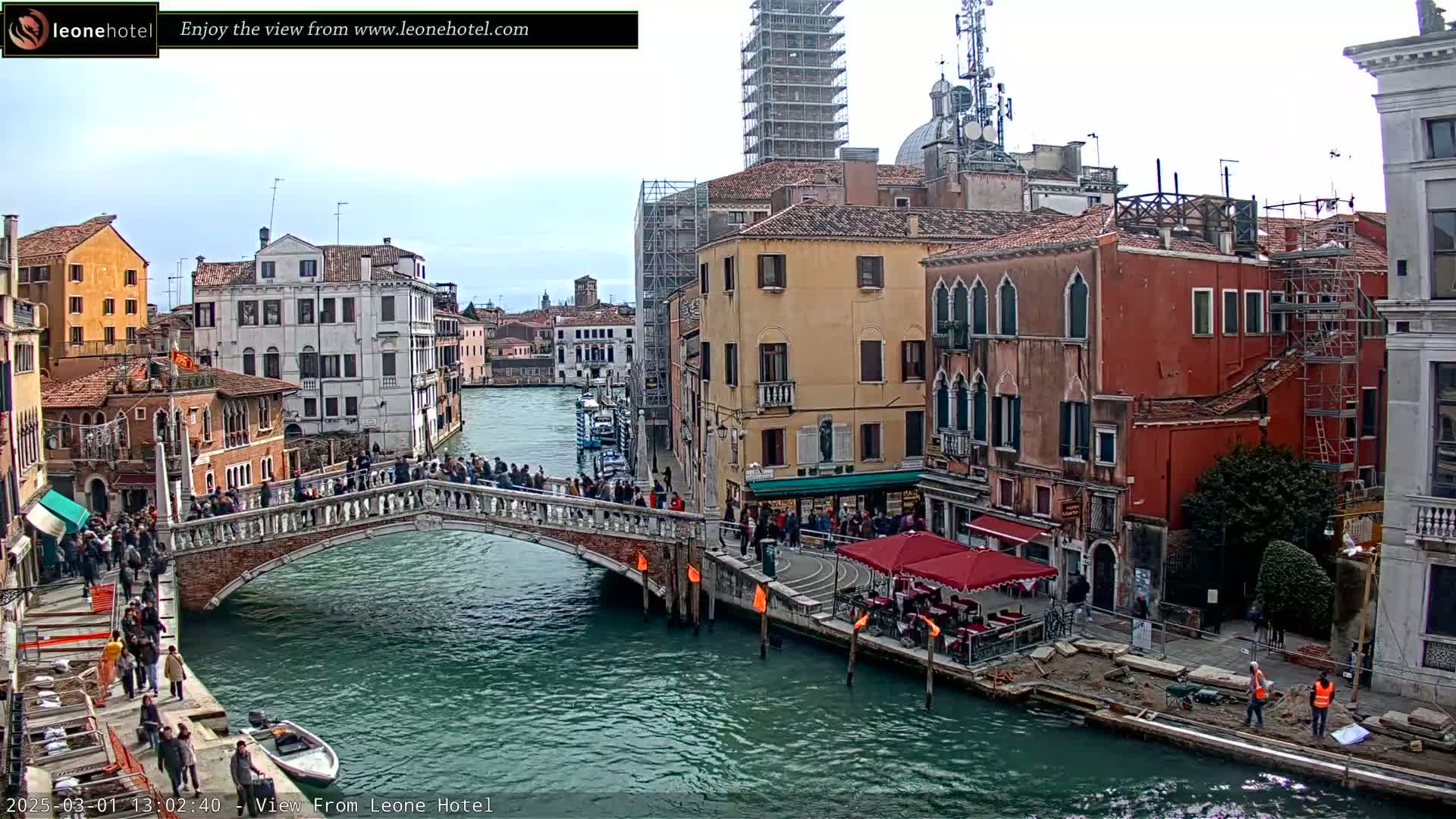 A bridge over a canal in Venice, Italy, is crowded with people under a mostly cloudy sky.