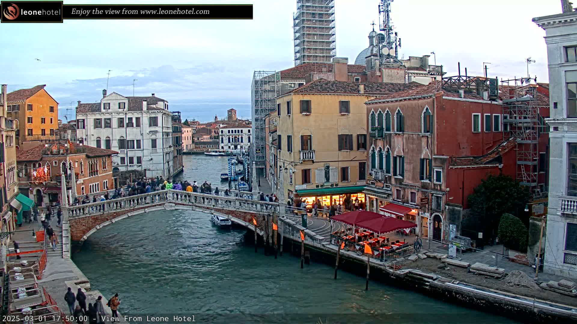 A bridge crosses a canal in Venice, Italy, under a cloudy sky, with numerous buildings lining the waterway and people walking on the bridge.