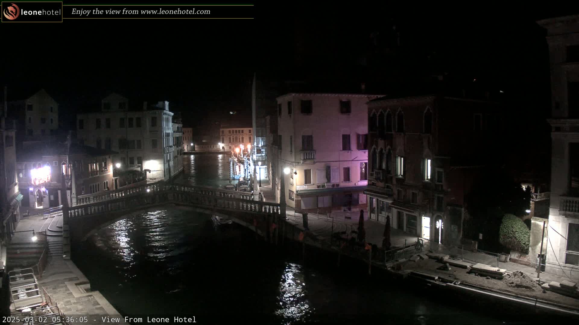A nighttime view of a canal in Venice, Italy, shows a stone bridge, buildings lining the waterway, and calm water reflecting lights.