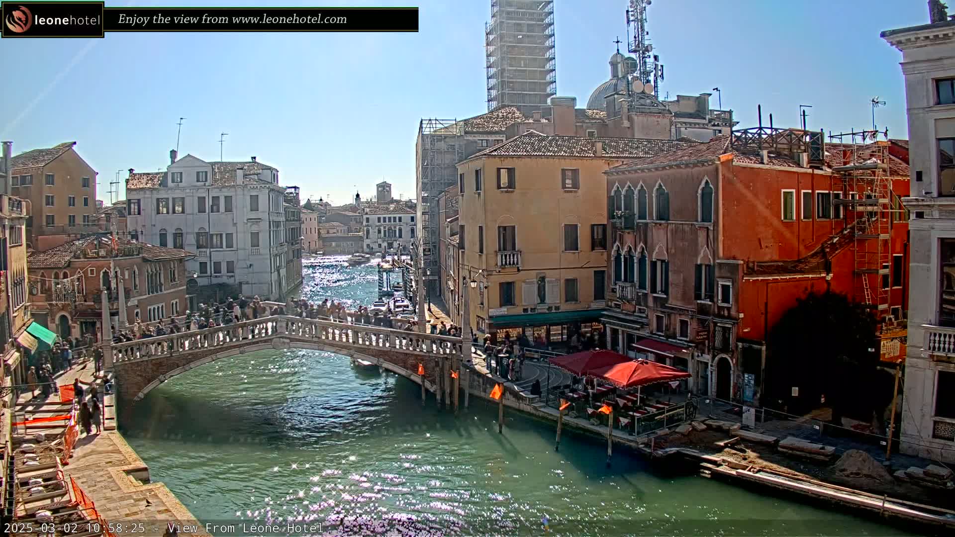A sunny day in Venice shows a bridge over a canal lined with buildings, some undergoing construction, with people walking across the bridge and along the canal.