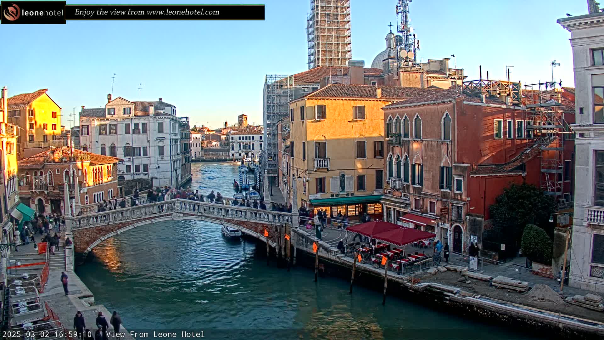 A stone bridge crosses a canal in Venice, Italy, under a clear blue sky, with numerous buildings lining the waterway, some under construction, and people walking across the bridge.