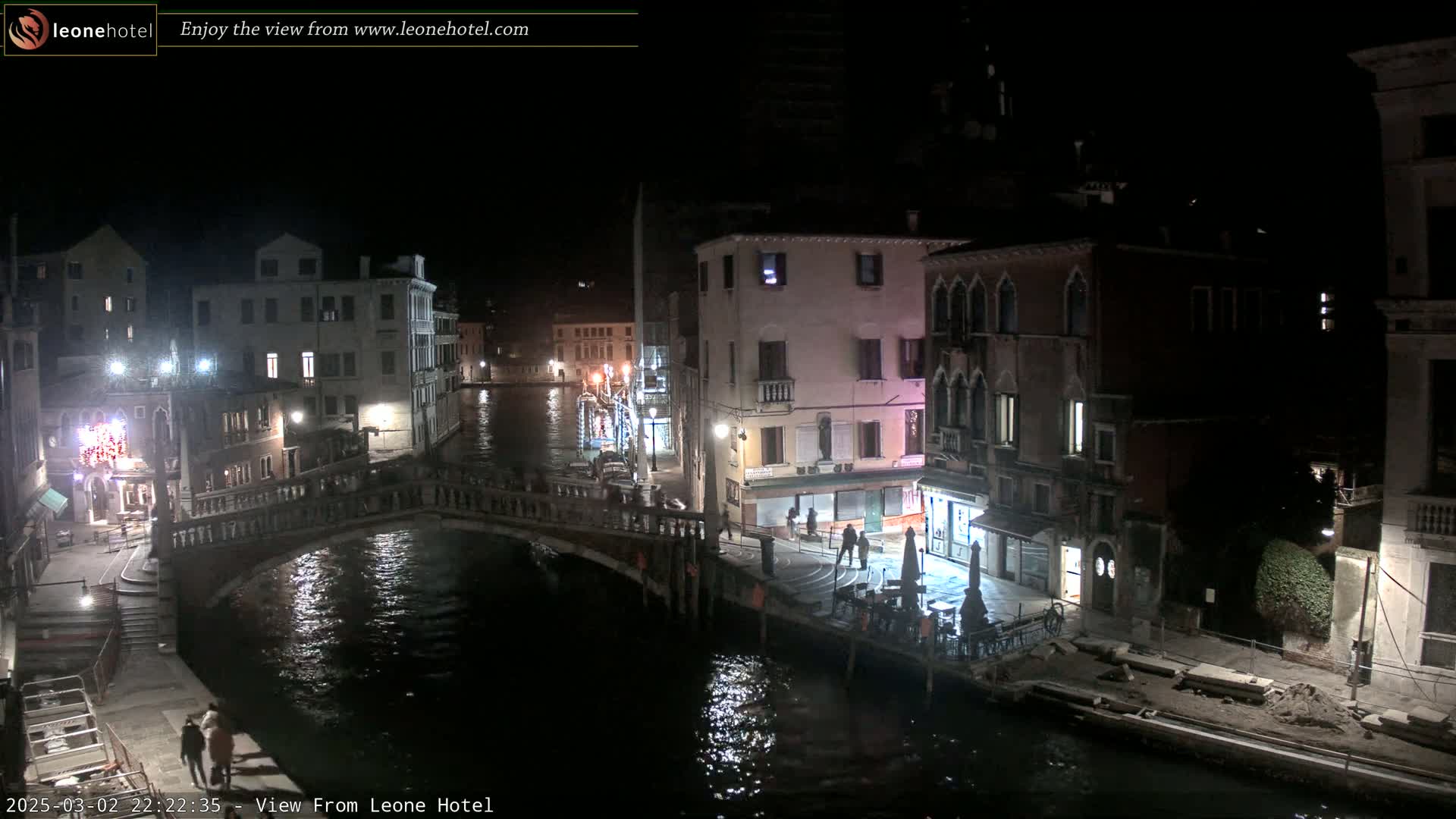 A nighttime view of a canal in Venice, Italy, shows several buildings illuminated along the waterway, a bridge crossing the canal, and a few pedestrians.