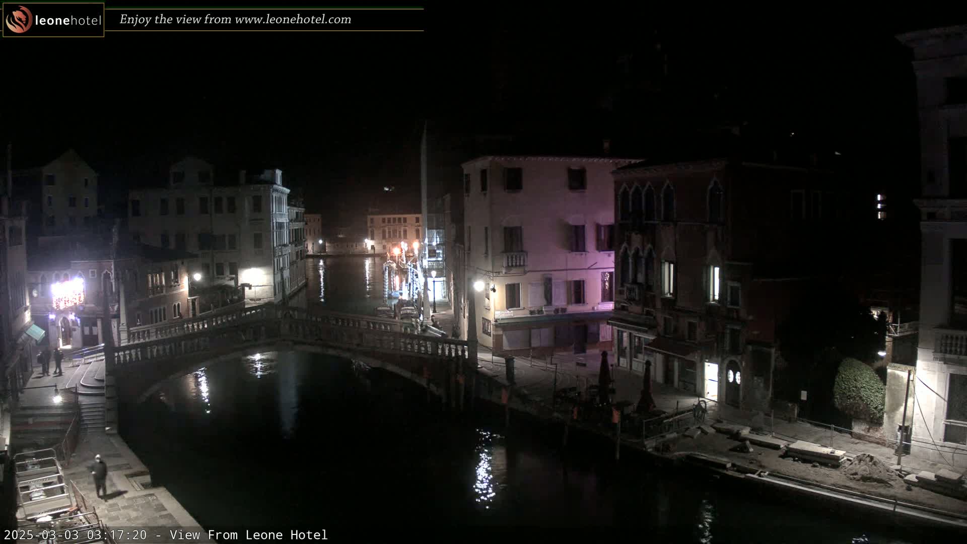 A nighttime view of a canal in Venice, Italy, shows several buildings lining the waterway and a bridge spanning the canal.